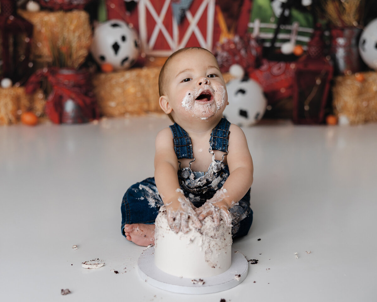 Little boy in blue overalls enjoying his cake smash, sitting on a white floor in a farm-style studio setup. Photograph by Yucaipa cake smash photographer Kaitlyn Dawn Photography.
