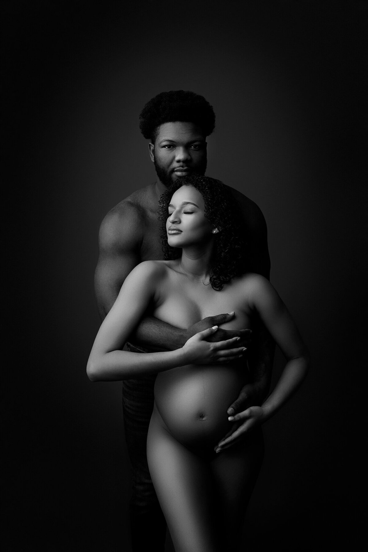 In this captivating black and white fine art maternity photograph, New Jersey's premier maternity photographer, Katie Marshall, skillfully frames a moment of intimate connection. The image features a woman facing the camera, her partner standing shirtless behind her. The man's forearm gently shields her breasts, while her head is turned to the left with closed eyes and a contemplative expression. Her hand cradles her baby bump with tenderness, and the man gazes at the camera with a serene, closed-mouth smile, revealing the depth of their connection in this timeless portrait.