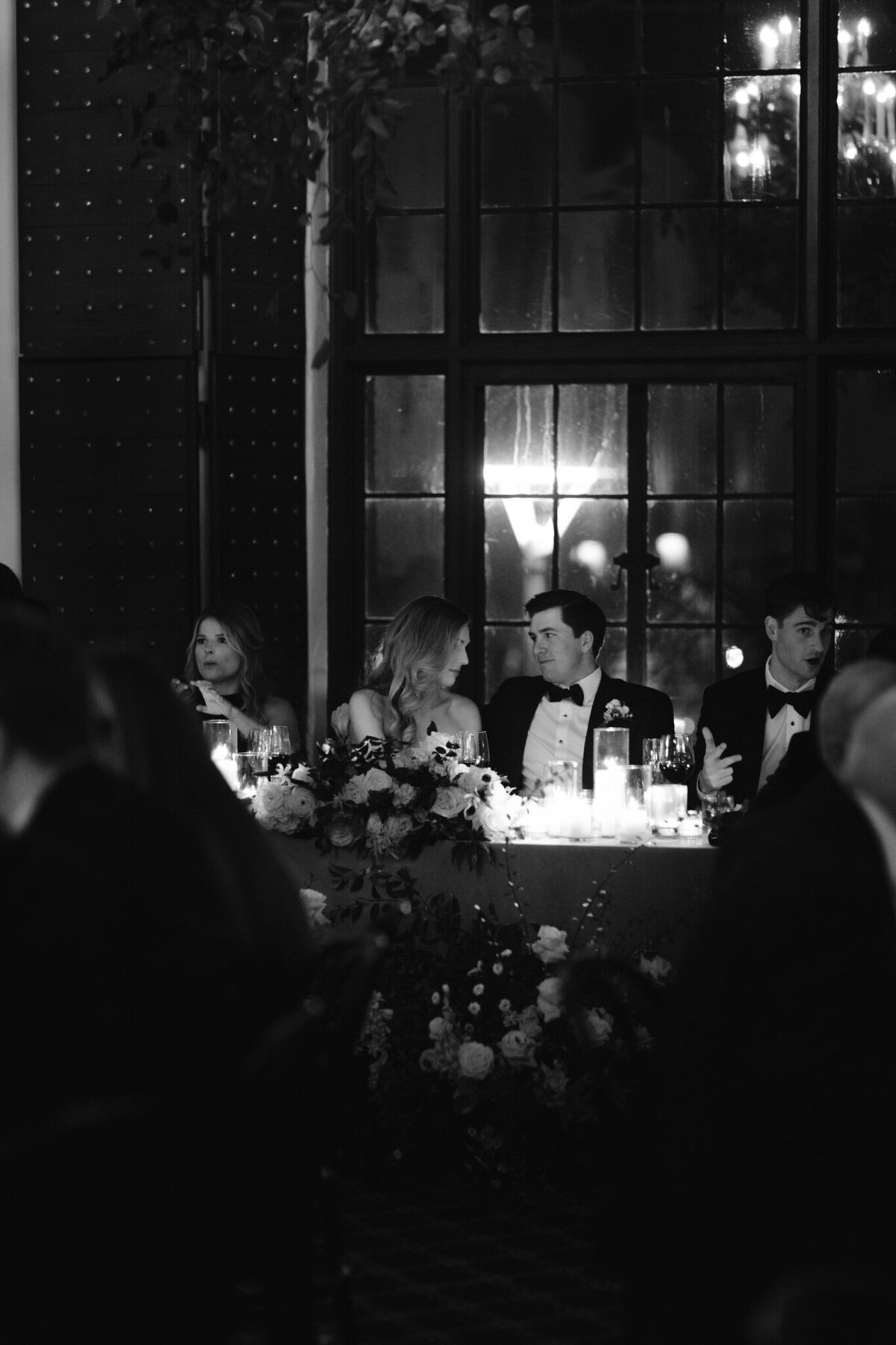 Black and white photo of wedding couple sitting at head table and talking to each other during wedding reception