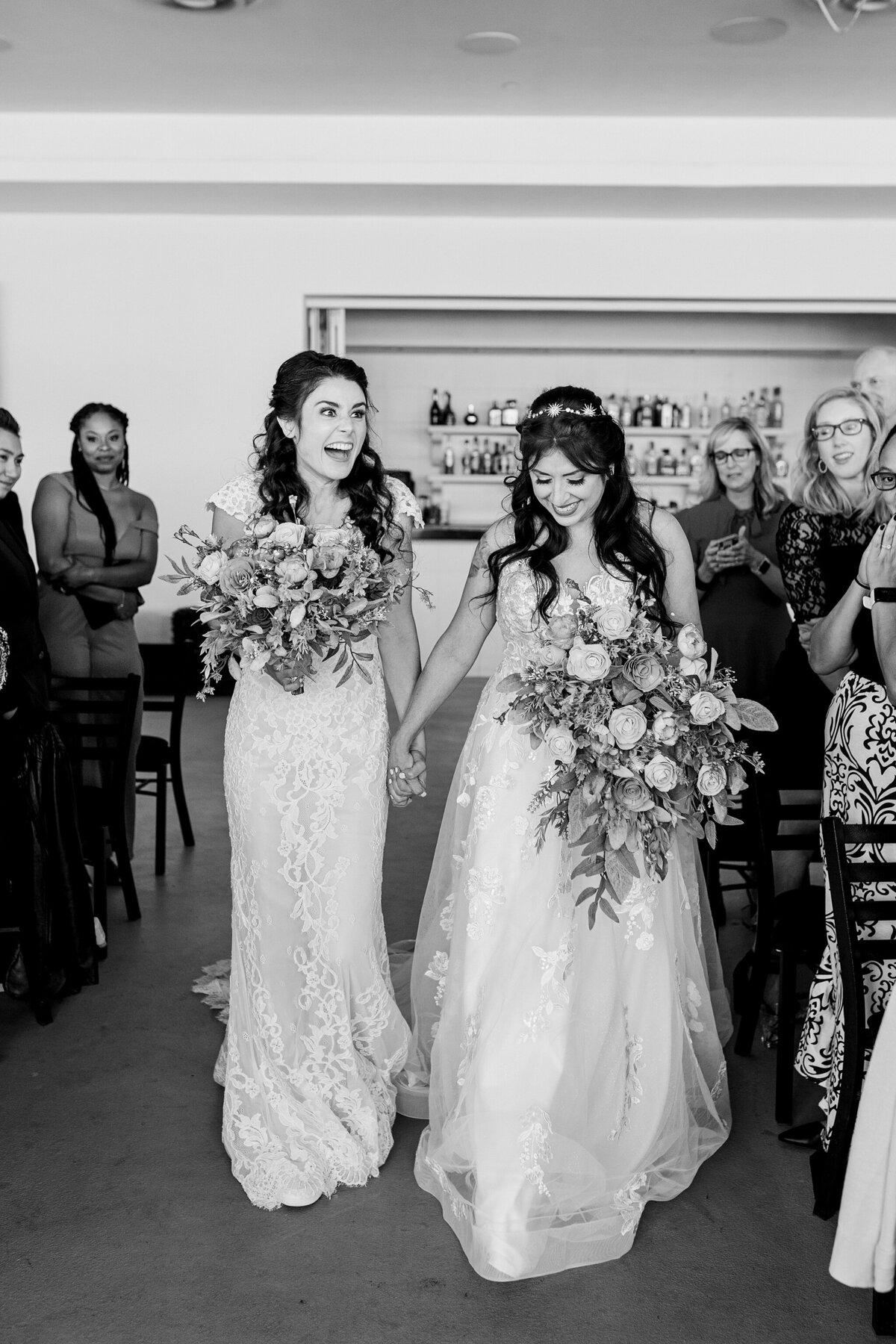 A candid photo of a lesbian couple walking down the aisle of their wedding ceremony at a wedding in Dallas, Texas. The bride on the left is heartily laughing, wearing a short sleeve intricate dress, and holding a bouquet. The bride on the right is looking down, smiling, is wearing a sleeveless intricate dress, and is holding a bouquet. Wedding guests can be seen watching on joyfully on either side of them.