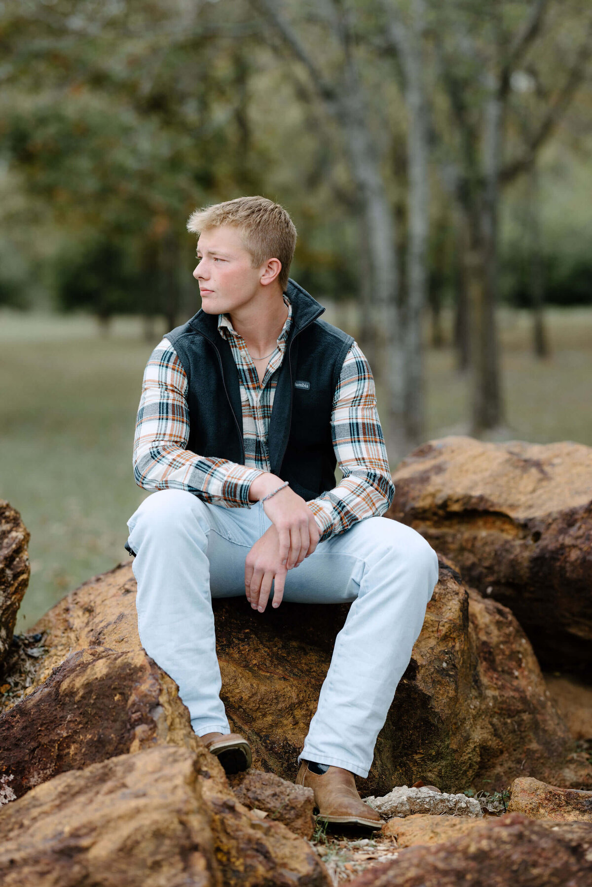senior boy in jeans, plaid flannel shirt, and black vest sitting in large rocks in Longview, TX park