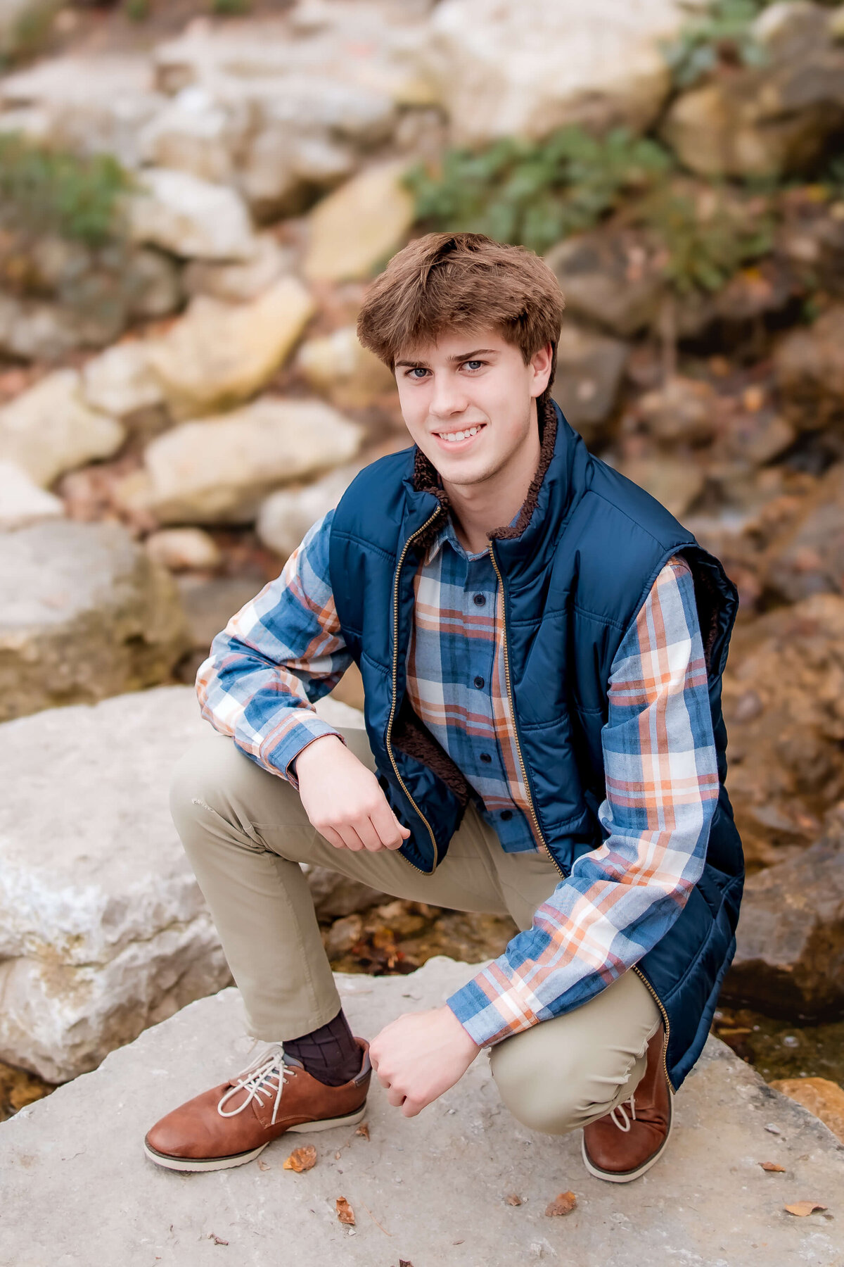 A cute young man is posing for senior photos on big boulders in the woods.