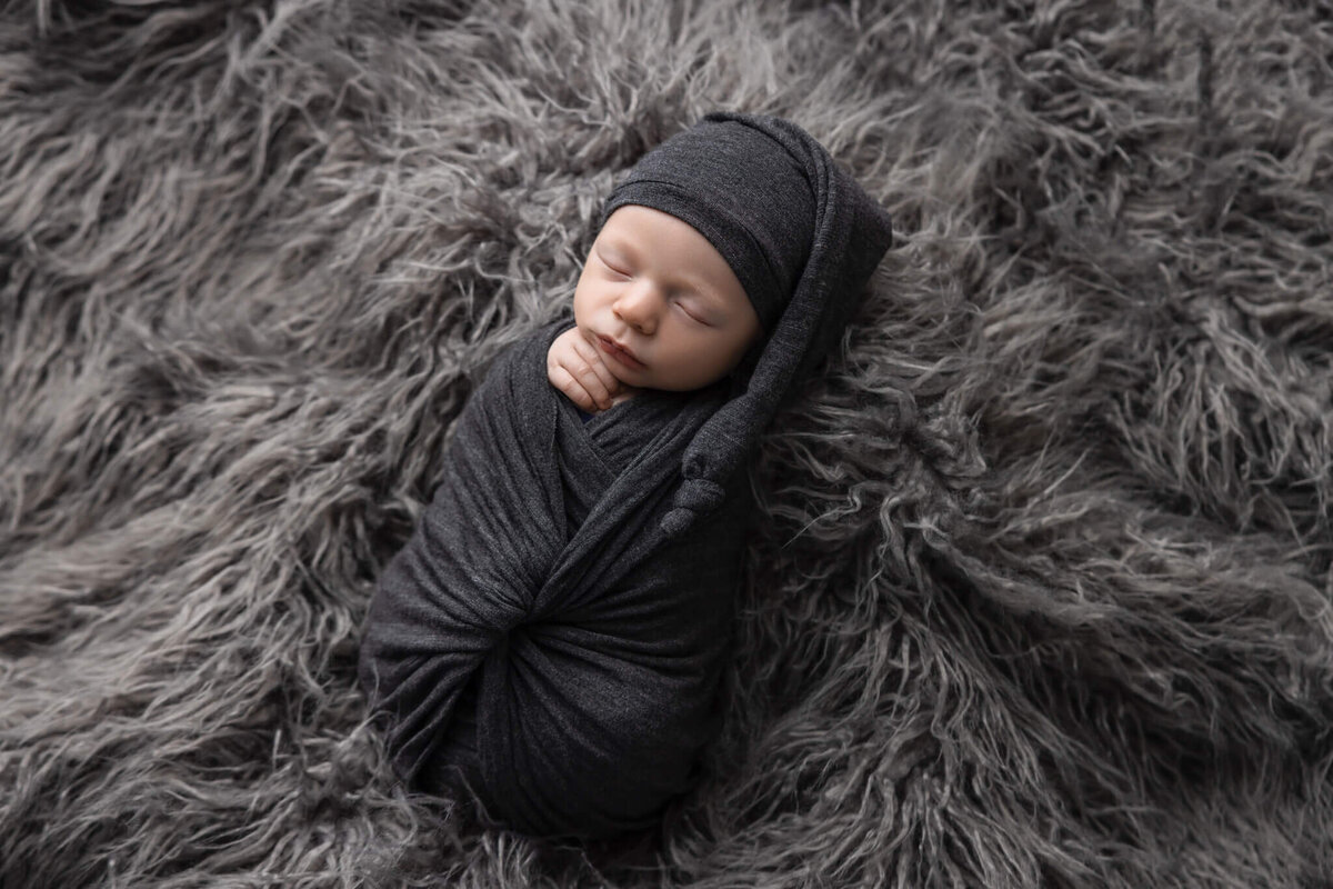 newborn baby asleep on grey shaggy rug wrapped in grey fabric wearing a grey sleepy hat