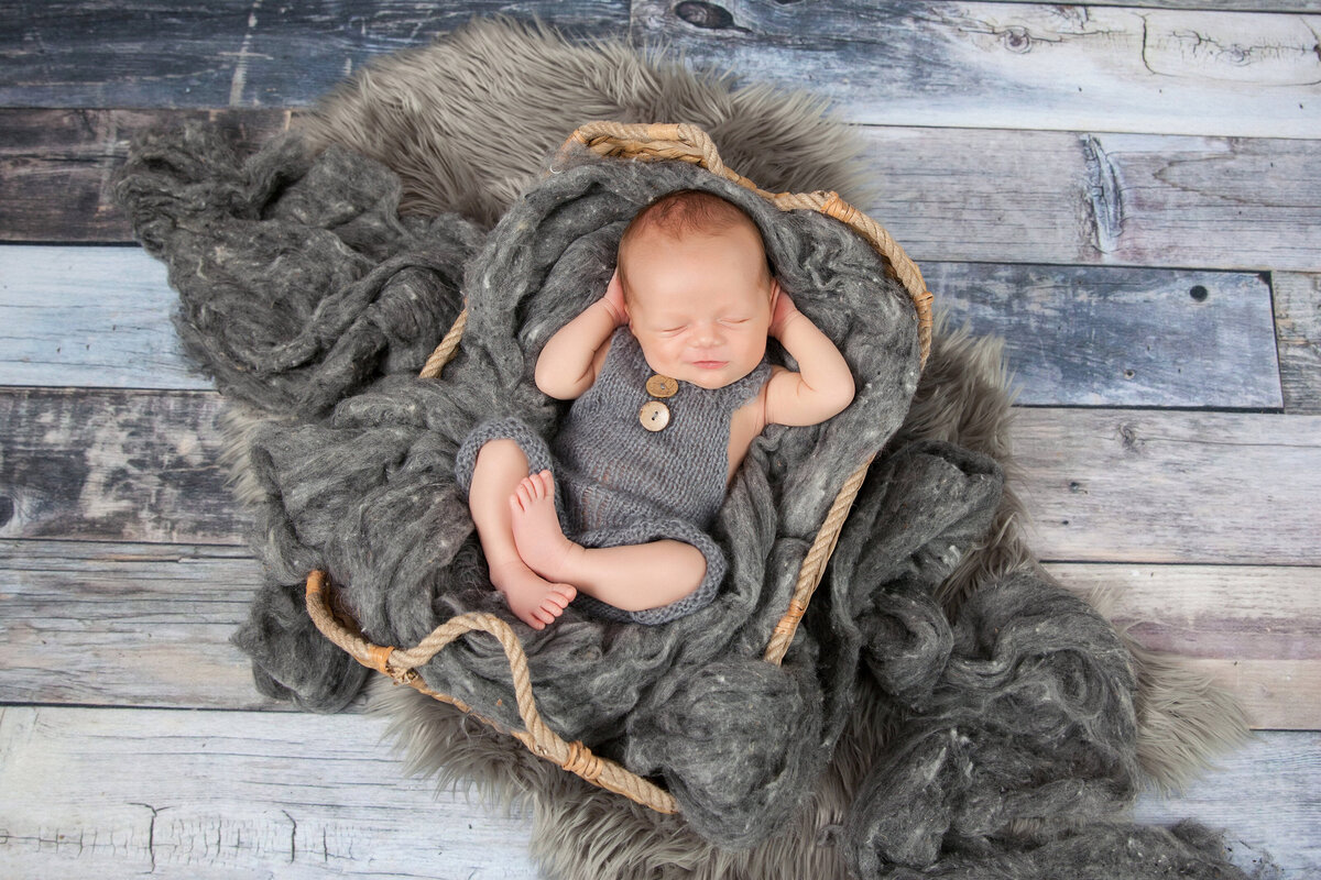 A baby sleeps peacefully in a basket lined with grey, fluffy fabric on a rustic wooden floor. Wearing a grey knitted romper, the little one has arms raised near the head, creating a serene scene captured by a Jacksonville baby photographer.