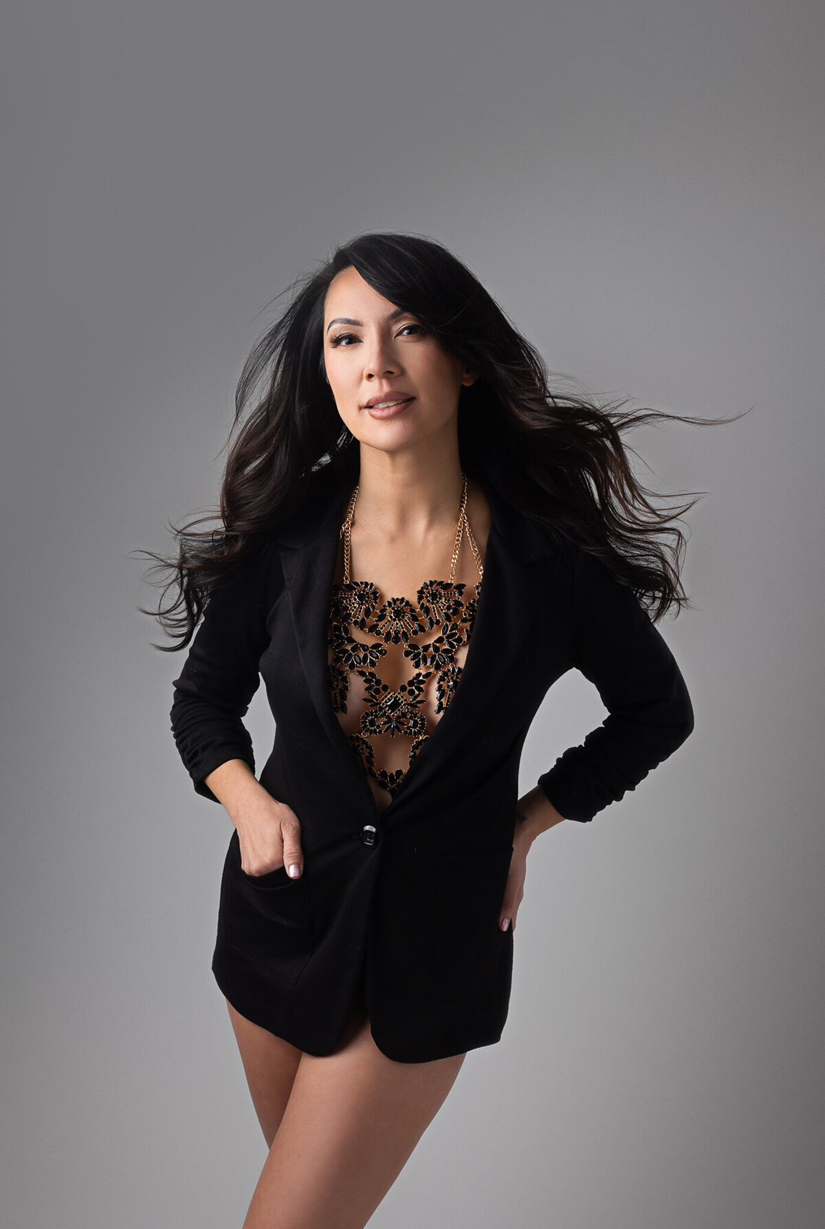 asian mature woman is posing with a blazer and a metal chain  white topless over a grey backdrop for her boudoir session