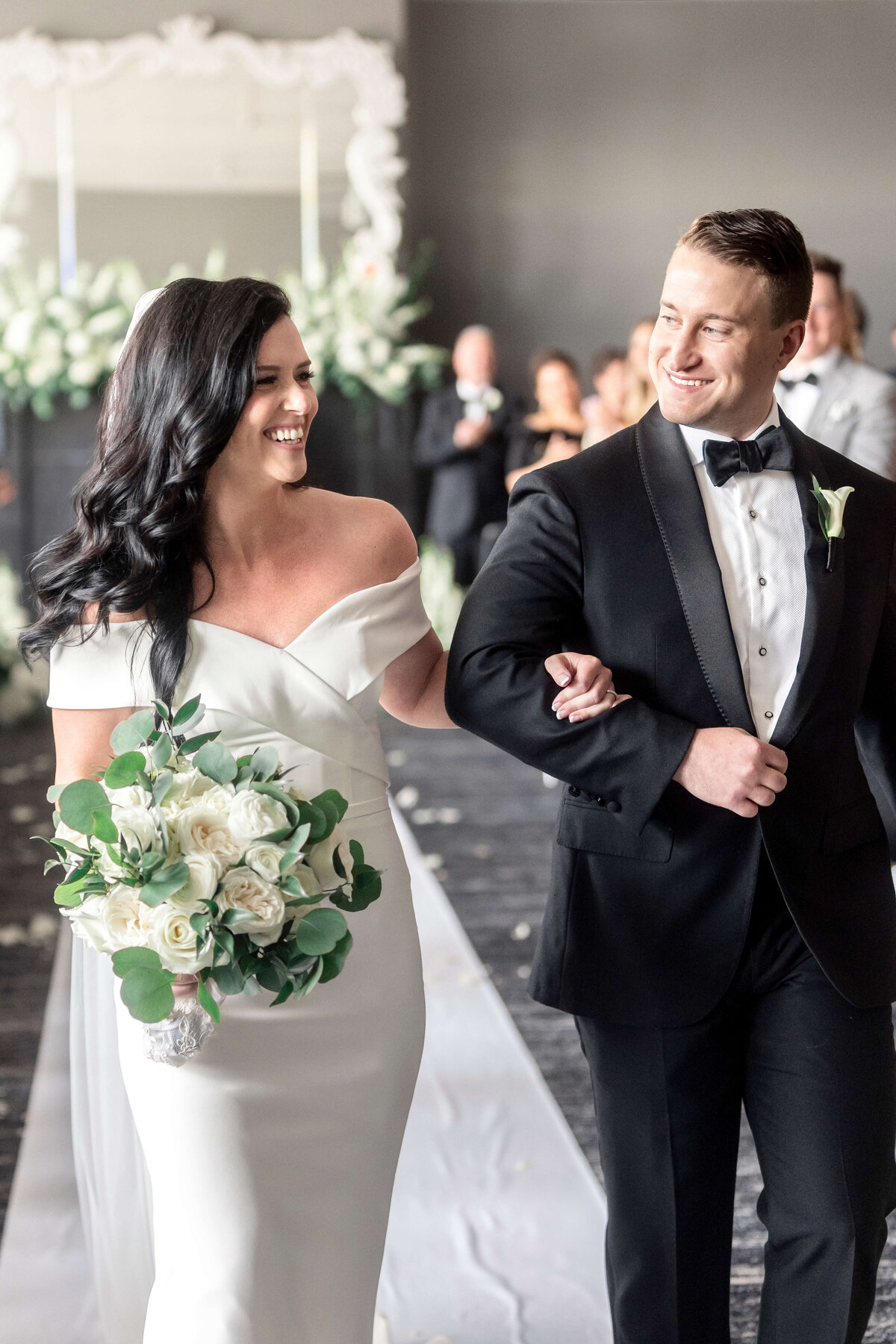 A bride in a white dress and groom in a black tuxedo walk arm in arm down an aisle. The bride holds a bouquet of white flowers and greenery. They are smiling at each other, with blurred guests and floral arrangements in the background.