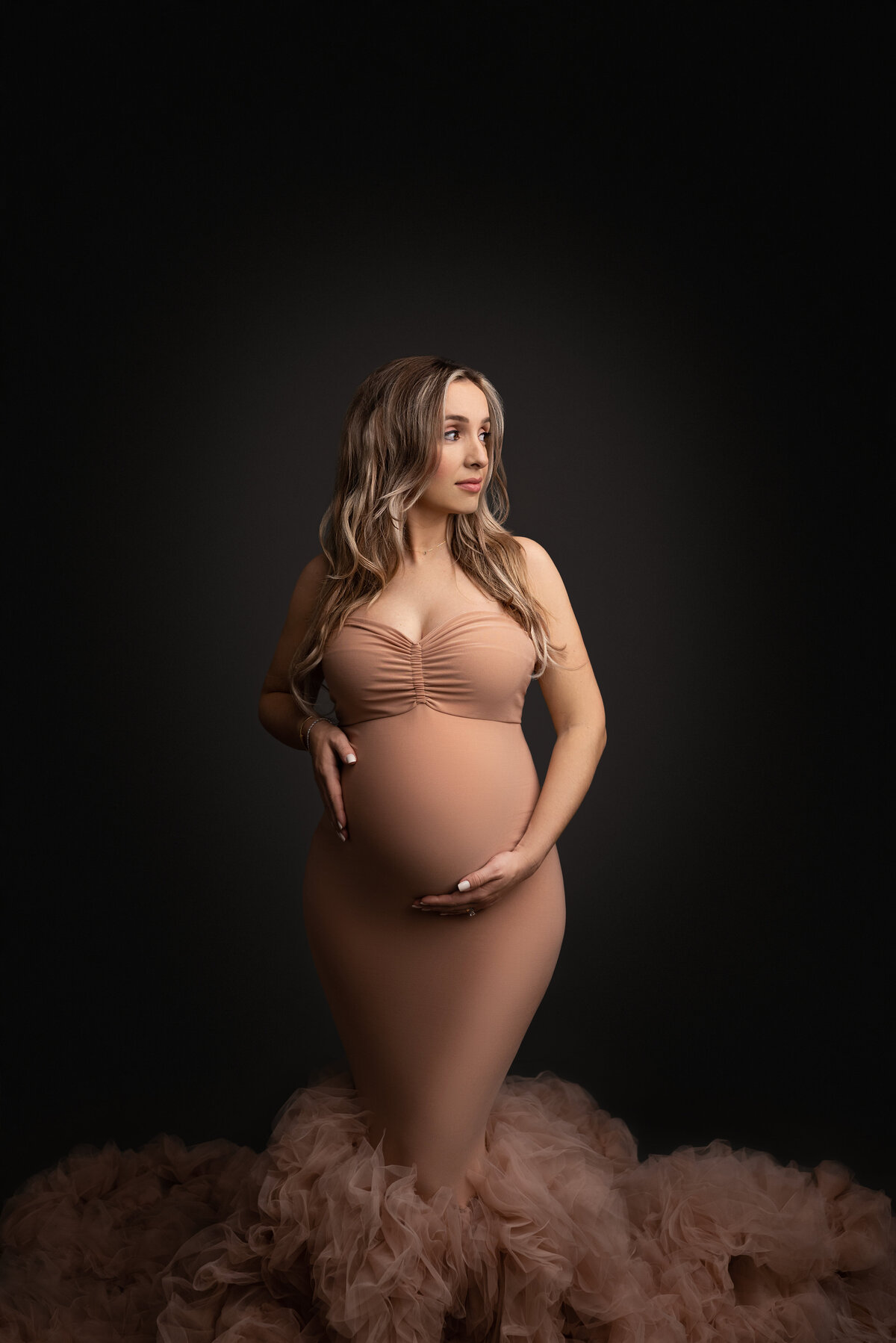Maternity photo taken by Katie Marshall, recognized as the best Main Line maternity photographer. The image showcases a woman standing against a grey background, gracefully dressed in a dark dusky blush body-con maternity gown featuring tulle ruching at the bottom. One of her hands tenderly cradles her baby bump, while the other rests gently at her side. She gazes over her shoulder, looking toward the distant light source, creating a graceful and timeless maternity portrait.