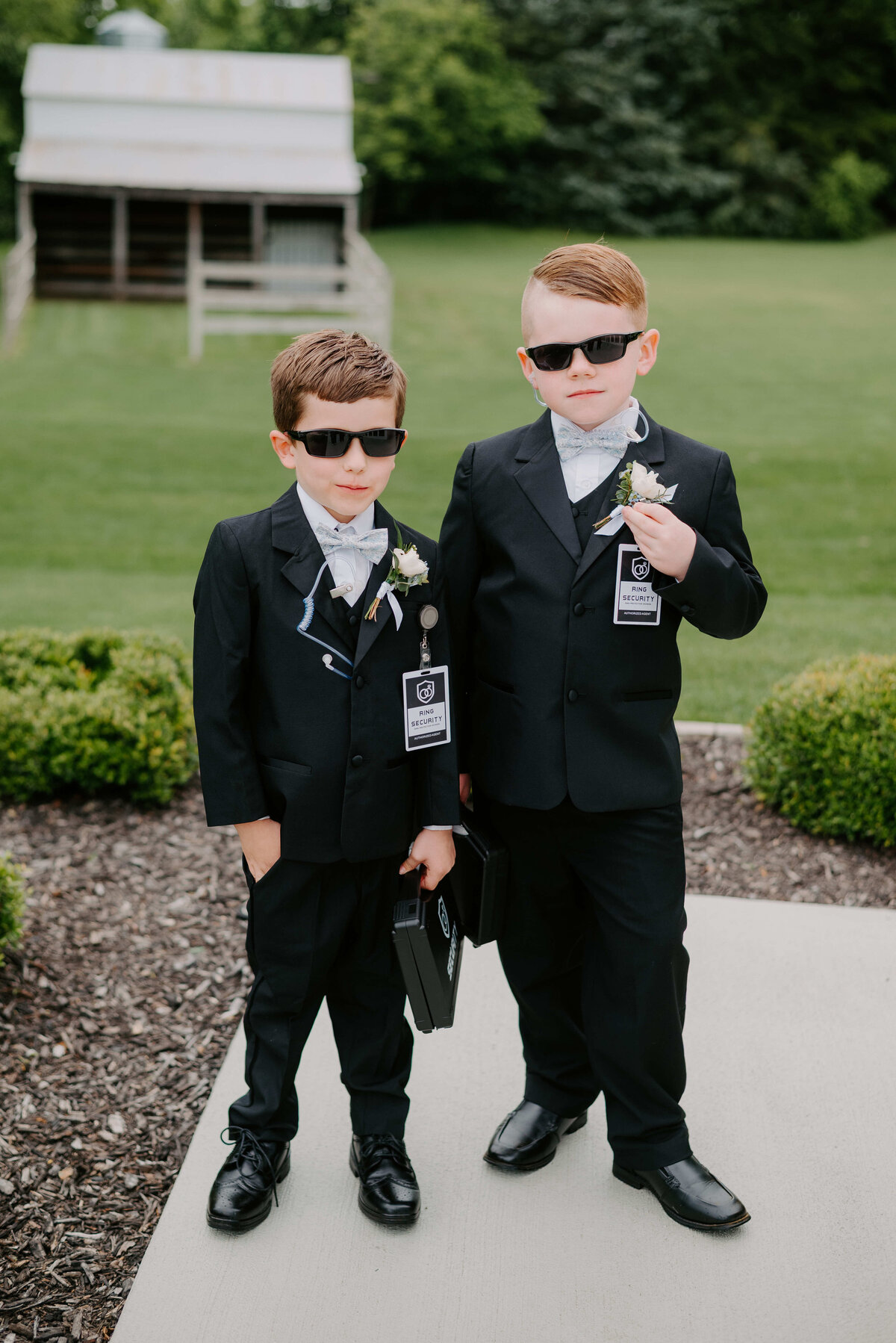 Two young boys in black suits and sunglasses stand confidently on a garden path. Each holds a small briefcase, creating a playful secret agent vibe.