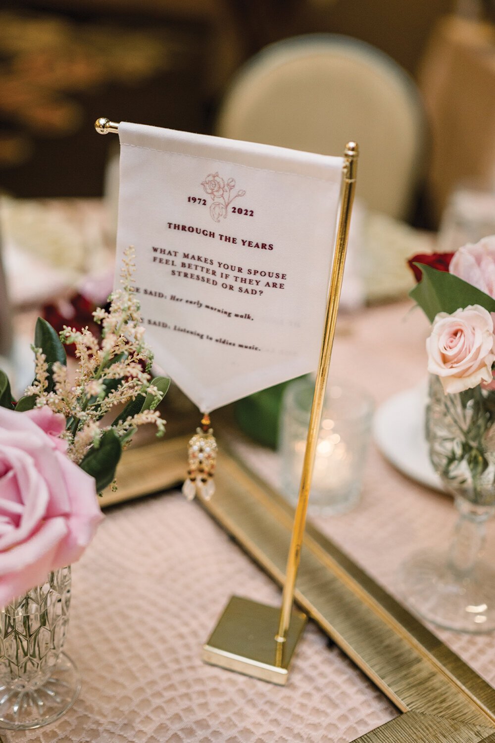 Table centerpiece for a 50th wedding anniversary featuring a custom fabric sign with beaded tassels hanging from a gold stand, surrounded by blush and burgundy florals in crystal vases, and a vintage gold frame.