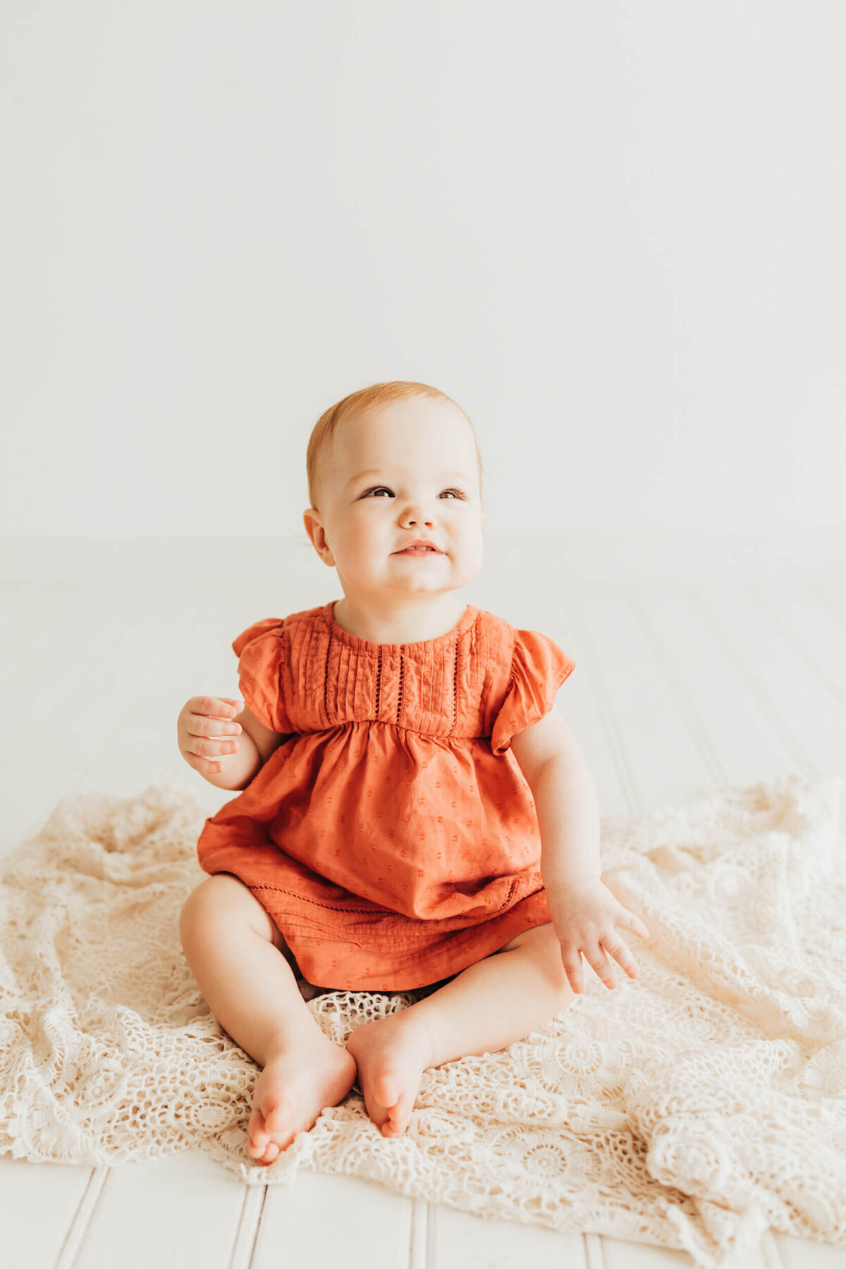 little girl wearing a rust colored dress, sitting on a crochet ivory blanket in studio with Allyson Blankenburg.
