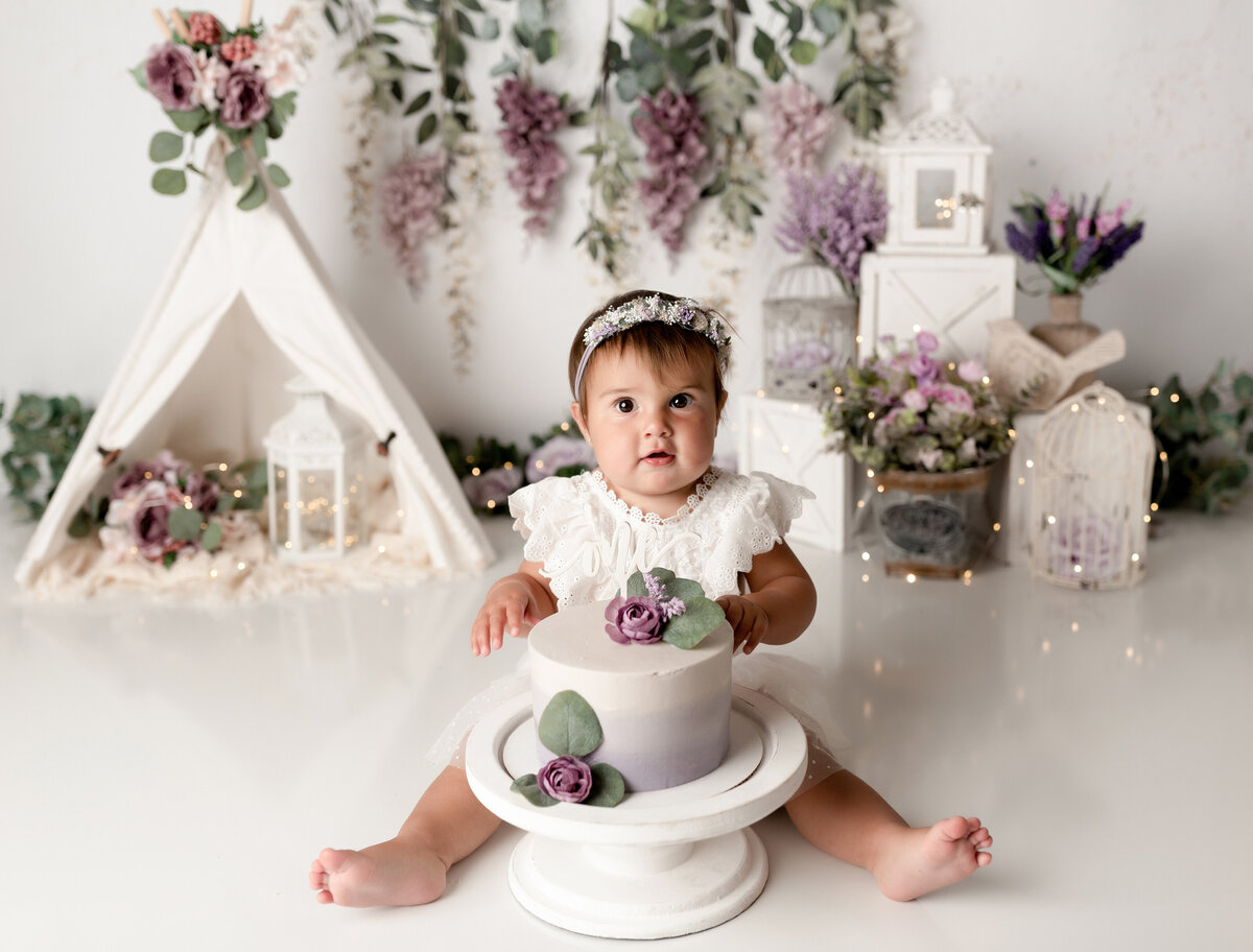 Lilac themed cake smash in West Palm Beach, FL. Baby girl is waring a white lace outfit sitting behind a purple ombre cake. She is looking at the camera about to touch the cake for the first time. The background has lilac flowers, a white canvas tent, and twinkle lights.