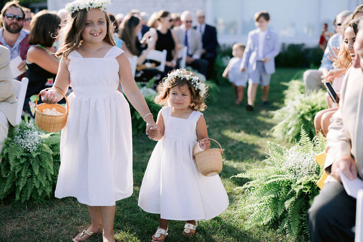 A little girl and an older girl are walking down the aisle in an outdoor wedding at Wianno, Cape Cod, Osterville, MA.