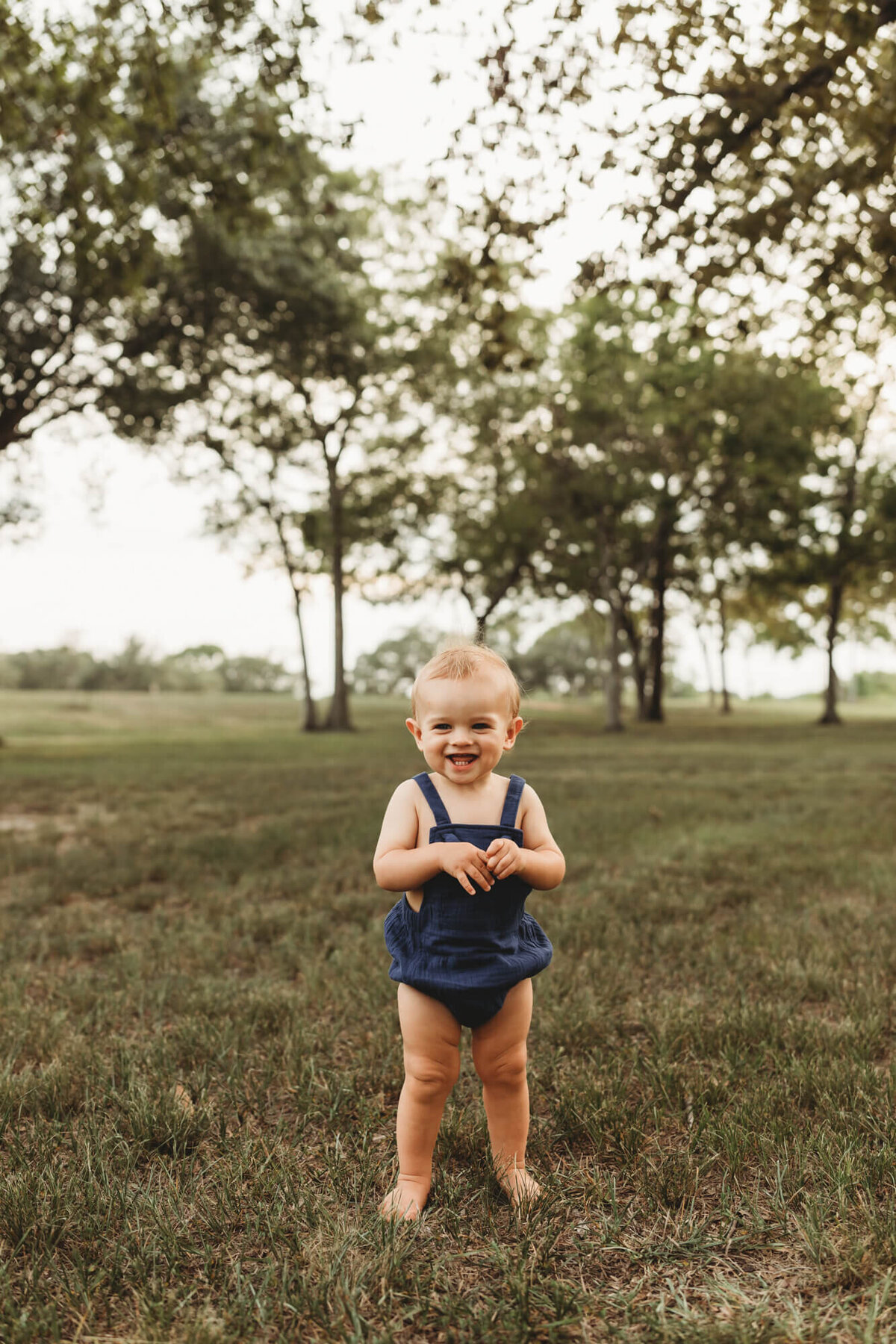 12 month old boy stands in blue romper on grass.