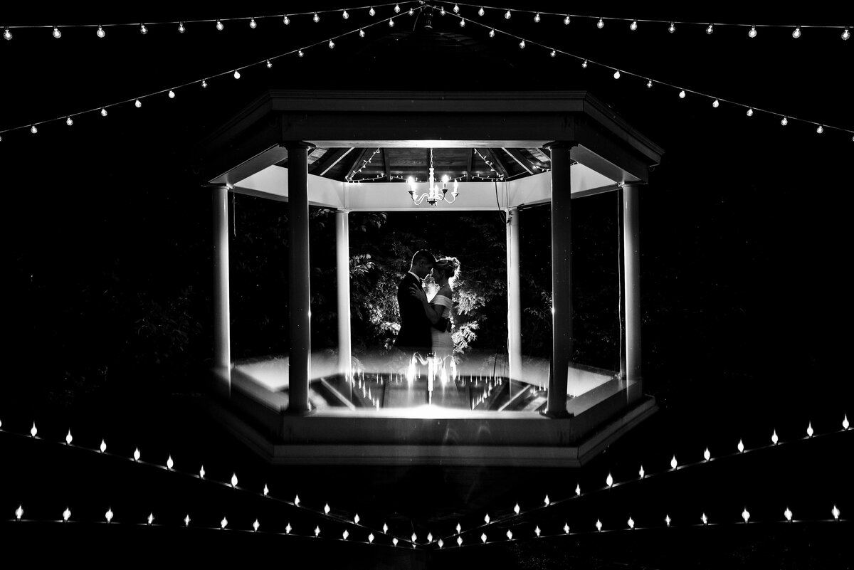 Black-and-white-image-of-a-rim-lit-silhouetted-couple-in-a-gazebo-at-night-with-fairy-light-and-the-top-of-the-gazebo-symmetrically-reflected-on-the-bottom-by-Charlotte-wedding-photographers-DeLong-Photography