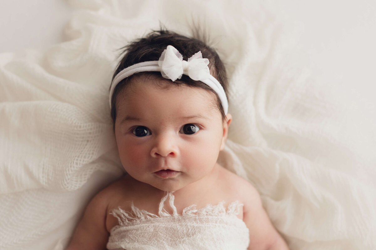 newborn baby girl with white bow looking at camera