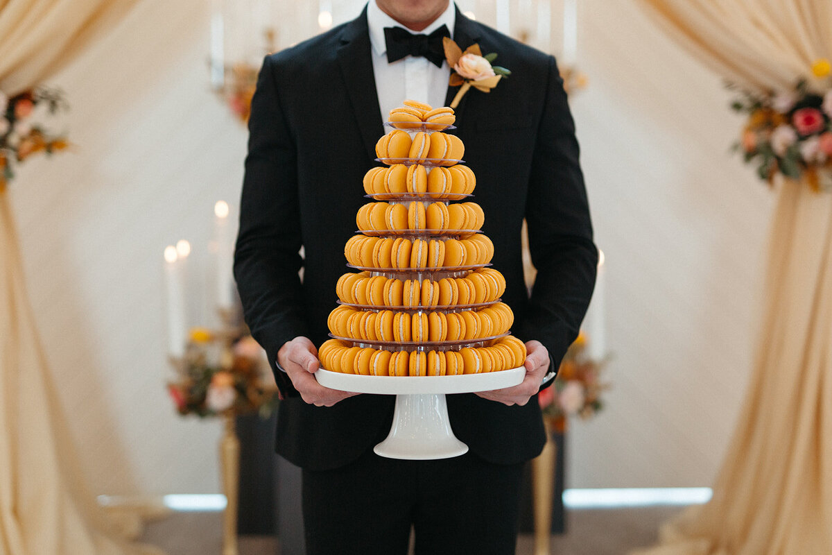 Close-up of the groom wearing a black tuxedo holding a cake stand filled with orange macaroons.