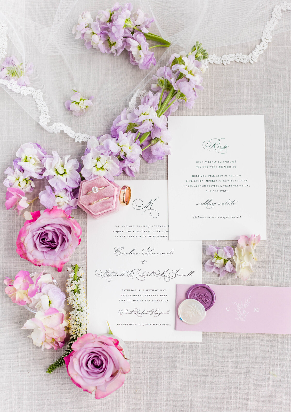 wedding invitation styled flat lay with pink ring box, purple and white florals and purple wax seals
