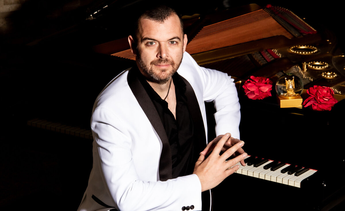 Classical musician portrait Jason Campbell wearing white dinner jacket leaning against piano keys beside red flowers