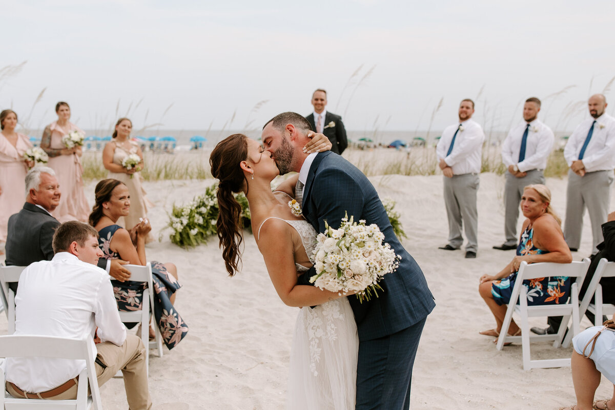 A  bride and groom kiss at the end of the aisle after just getting married. They are on a beach and their wedding guests are seated behind them