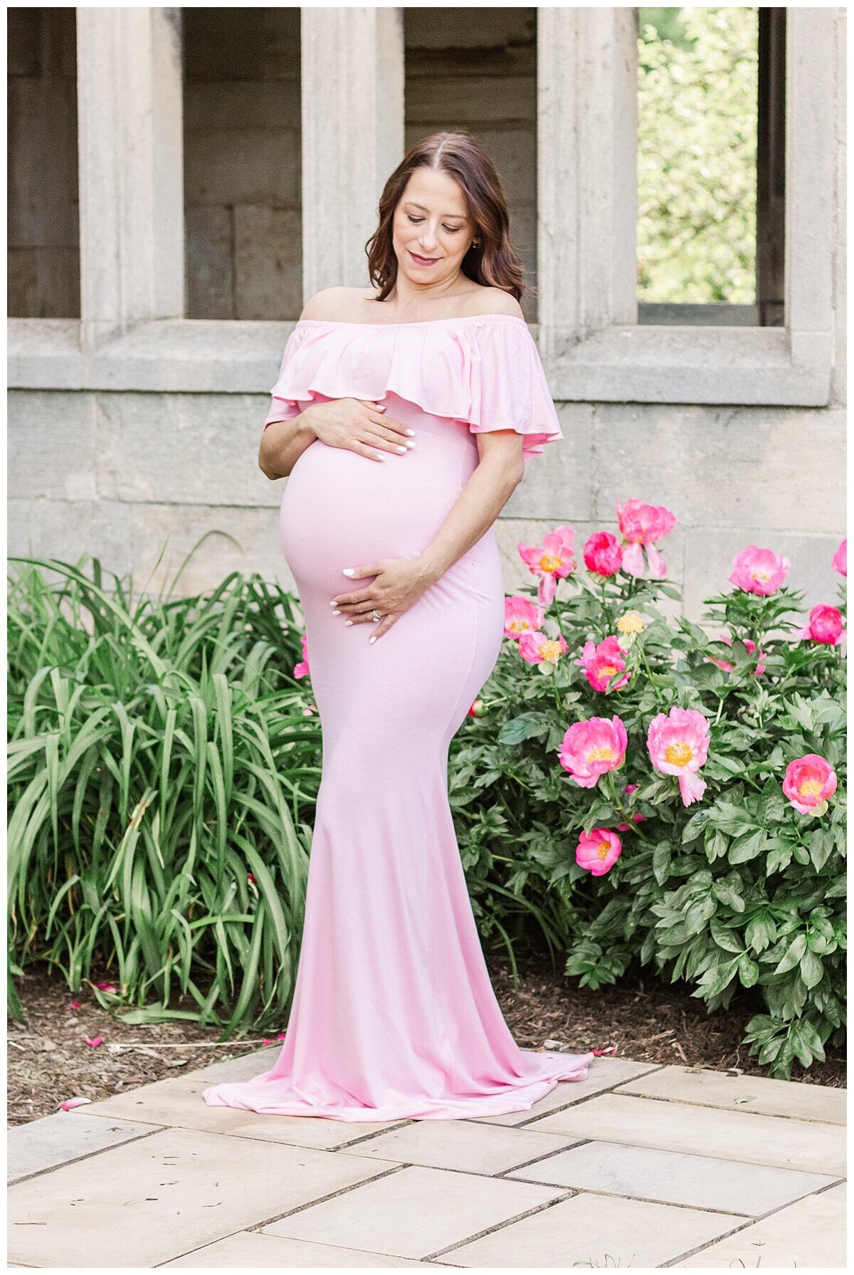 Maternity Photography in Wexford taken outdoors.  the mom is standing in front of pink flowers while wearing a pink dress.  she is also looking at her beautiful baby belly.