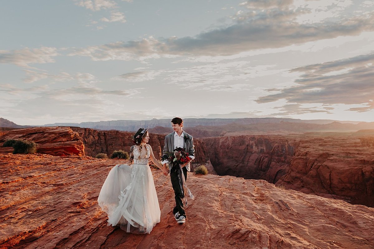 Groom and Bride walk hand in hand over orange desert overlooking horseshoe bend
