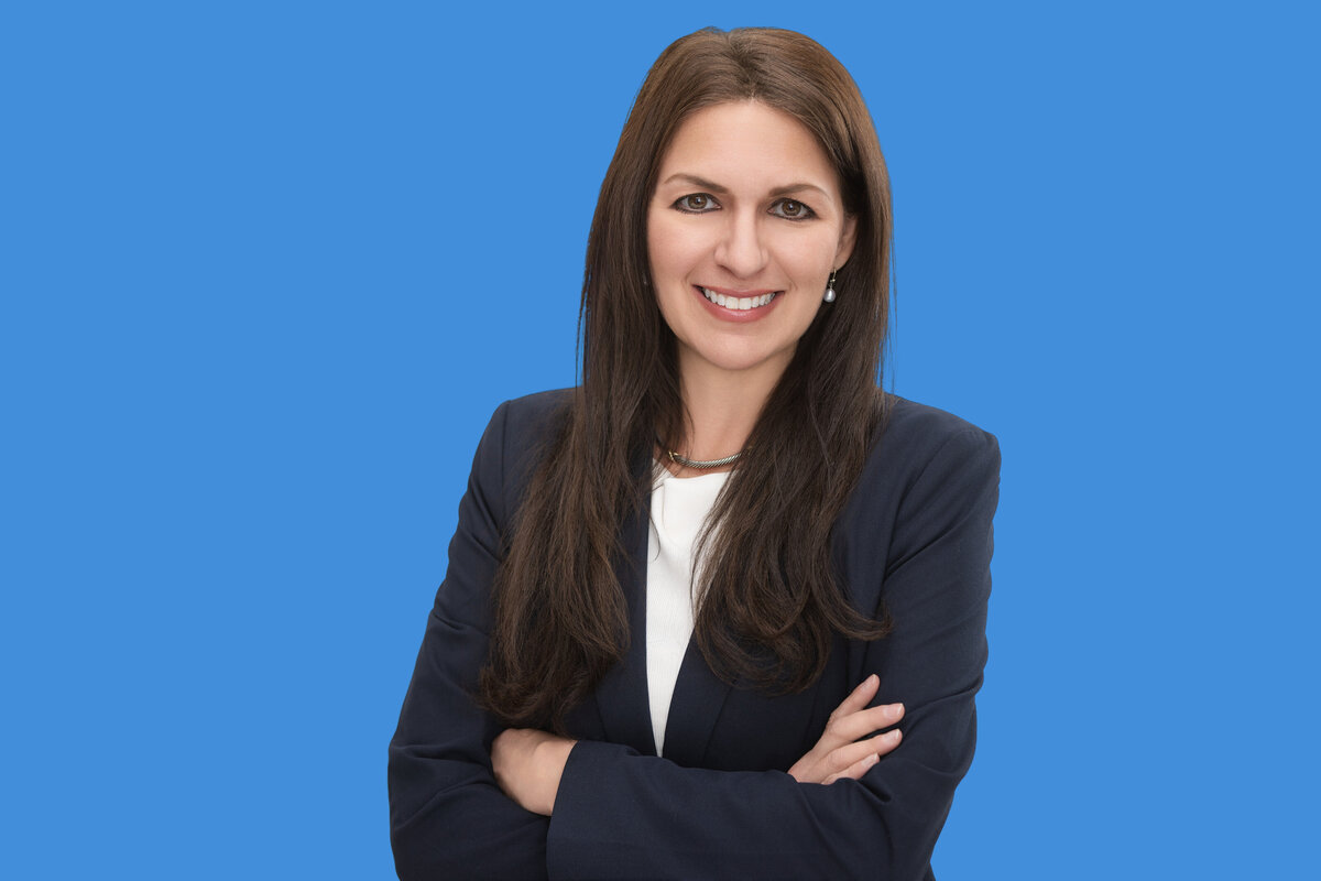 Clean and vibrant studio headshot of a brunette businesswoman with long straight hair, pearl drop earrings, and a confident smile, wearing a classic navy blazer and white blouse. Set against a bold blue background, this image was taken at Janel Lee Photography—Cincinnati’s trusted studio for corporate headshots, speaker branding portraits, and personal branding photography. Ideal for professionals in finance, legal, consulting, and sales roles looking to refresh their LinkedIn profile or business bio with a polished and approachable look.
