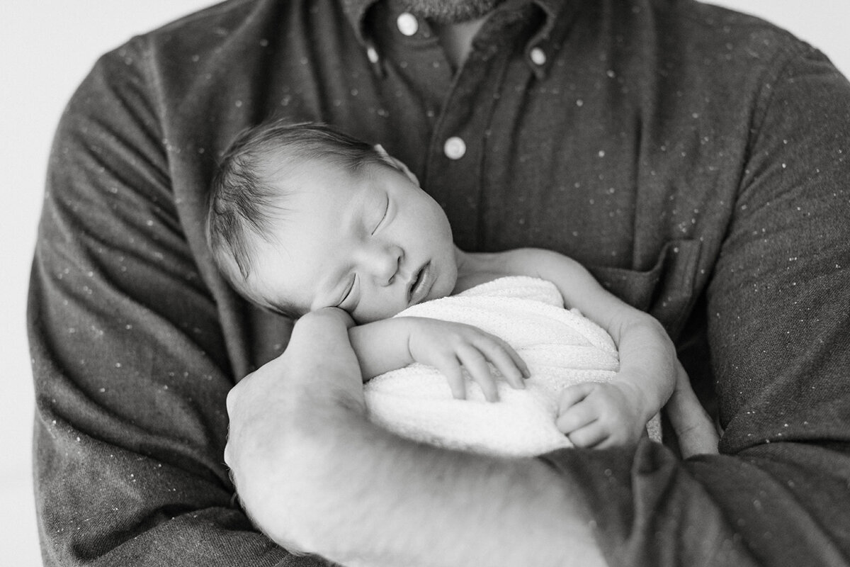 Newborn back is to dad’s stomach while dad’s arms wrap around baby and prop her up while she sleeps