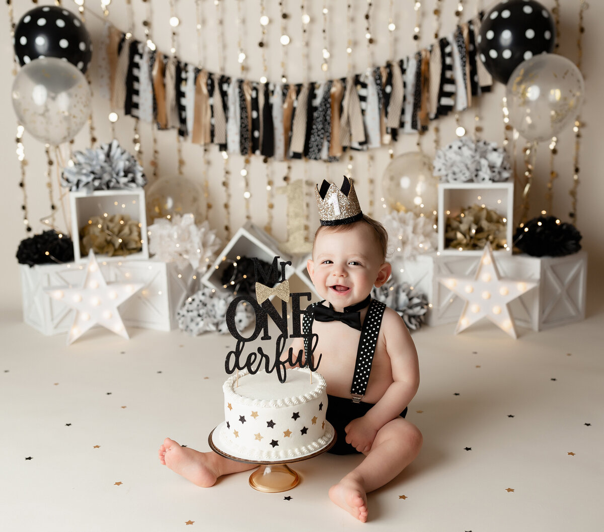 Black, white and gold cake smash at West Palm Beach and Wellington newborn and cake smash photographer. Baby boy in black shorts, black and white polkadot suspenders, and gold party crown sitting in front of a white cake with black and gold stars. In the background a gold white and black pennant, gold wall hangings, balloons, and white stars.