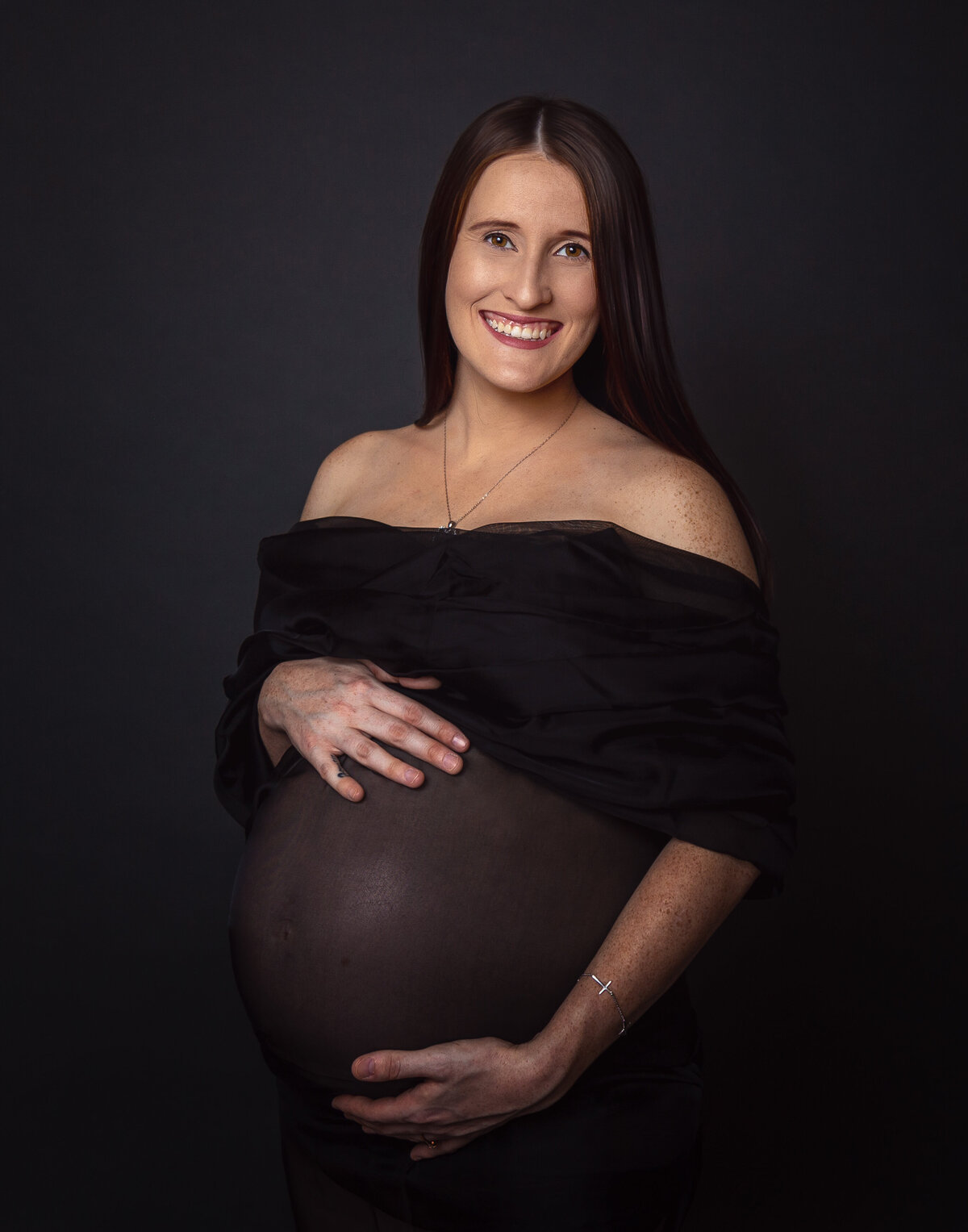 pregnant woman smiling in a see-through belly dress with right hand on belly and left hand under