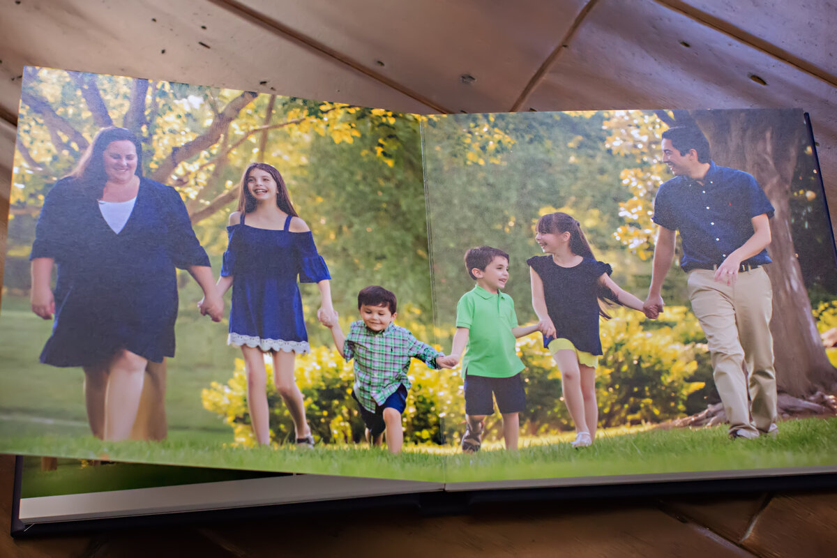 Photo Cover Session Book opened to a full bleed two page spread featuring a family of six with parents and four children holding hands and walking together, shown in a seamless lay flat design that highlights the storytelling flow and premium print quality.