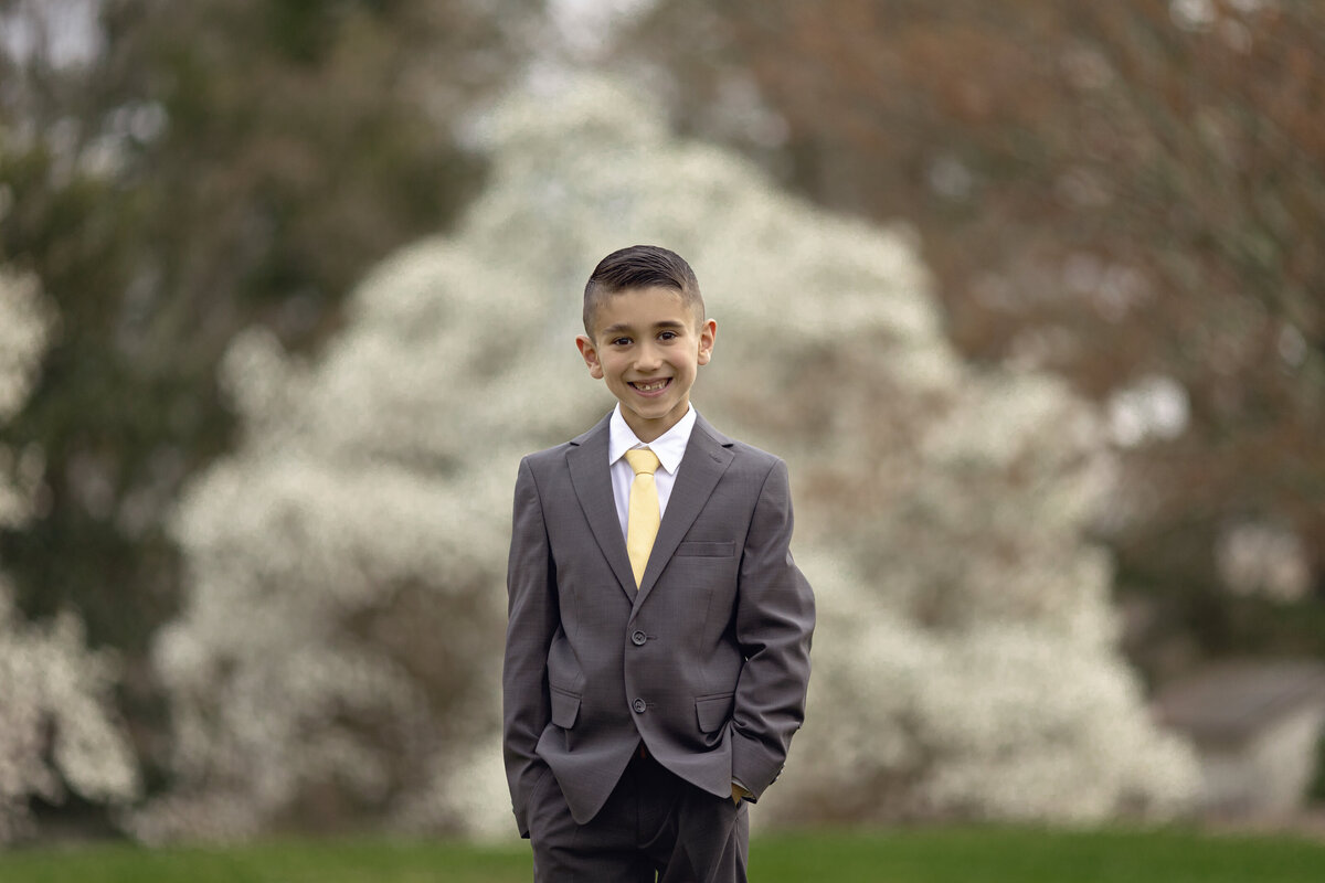 A boy in a grey suit smiles with hands in his pockets in front of a white flowering tree in a park