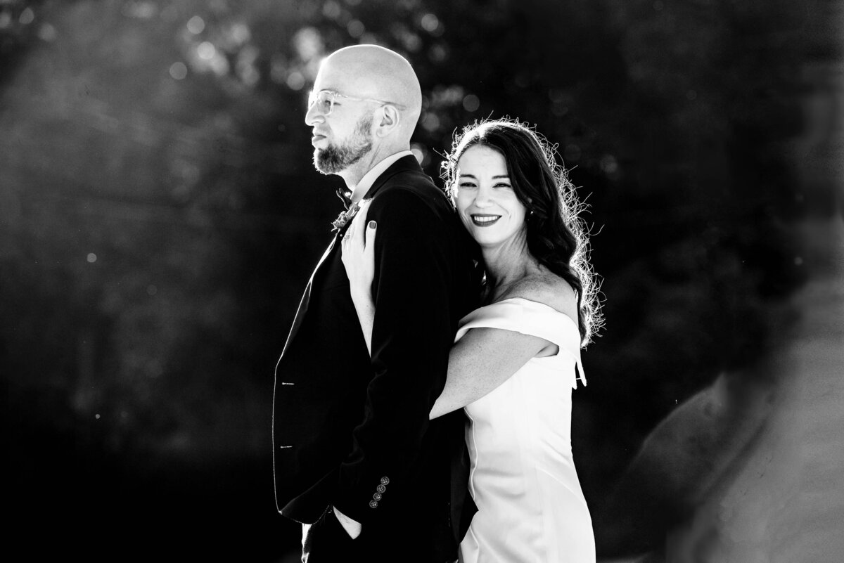 Black-and-white-photograph-of-a-bride-smiling-at-the-camera-as-she-hugs-her-groom-from-behind