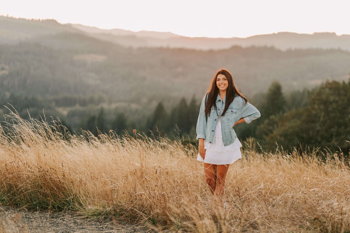 A high school senior girl stands in the grass at Fitton Green in Corvallis, OR. Her hand is on her hip and the mountains are in the background.