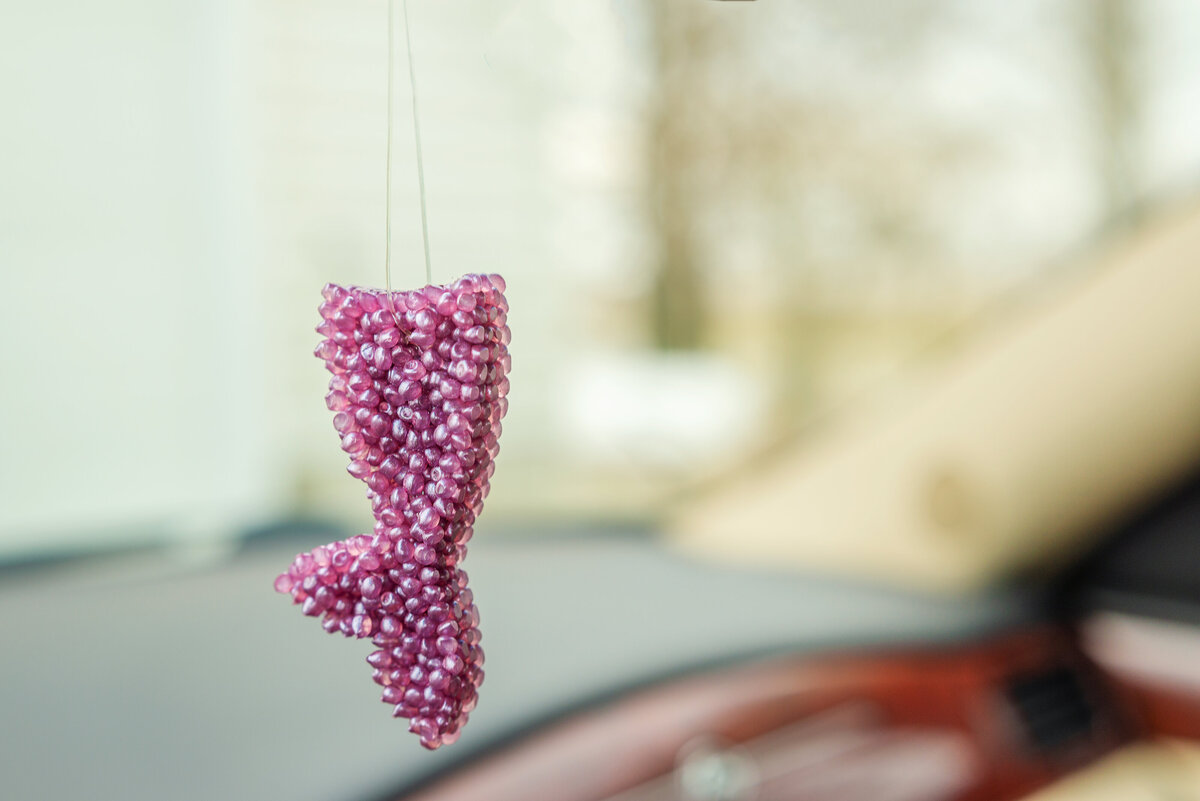 A pink beaded car air freshener in the shape of a dress, hanging from a car's rearview mirror with a blurry background.