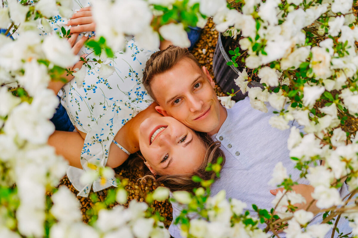 Photo from overhead of a man and woman laying on each other shoulder surrounded by flowers