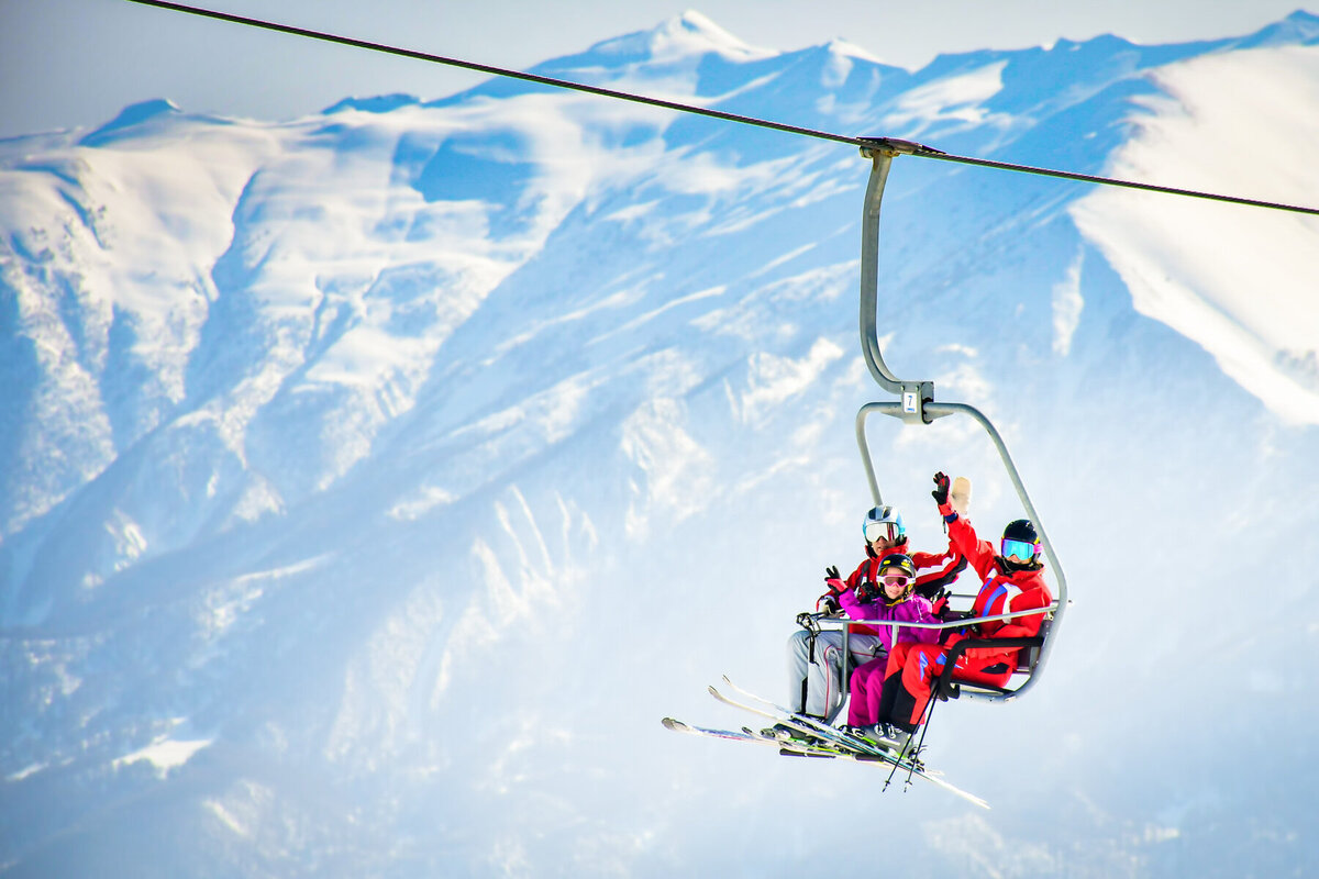Four people in colorful ski gear are riding a chairlift over snowy mountains. The background features expansive snow-covered peaks under a clear blue sky. One person waves, adding a sense of excitement to the scene.