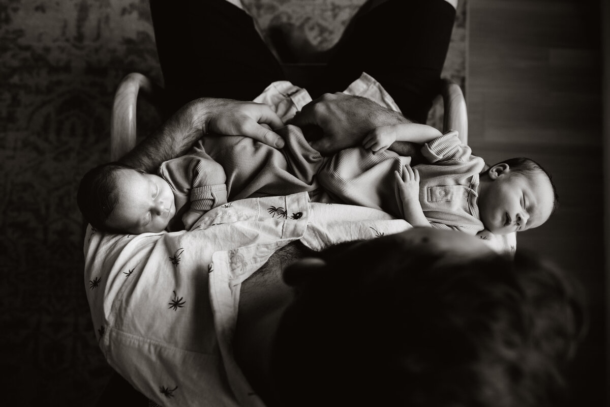 black and white portrait from above with a  man sitting in a chair holding twin baby boys while they sleep