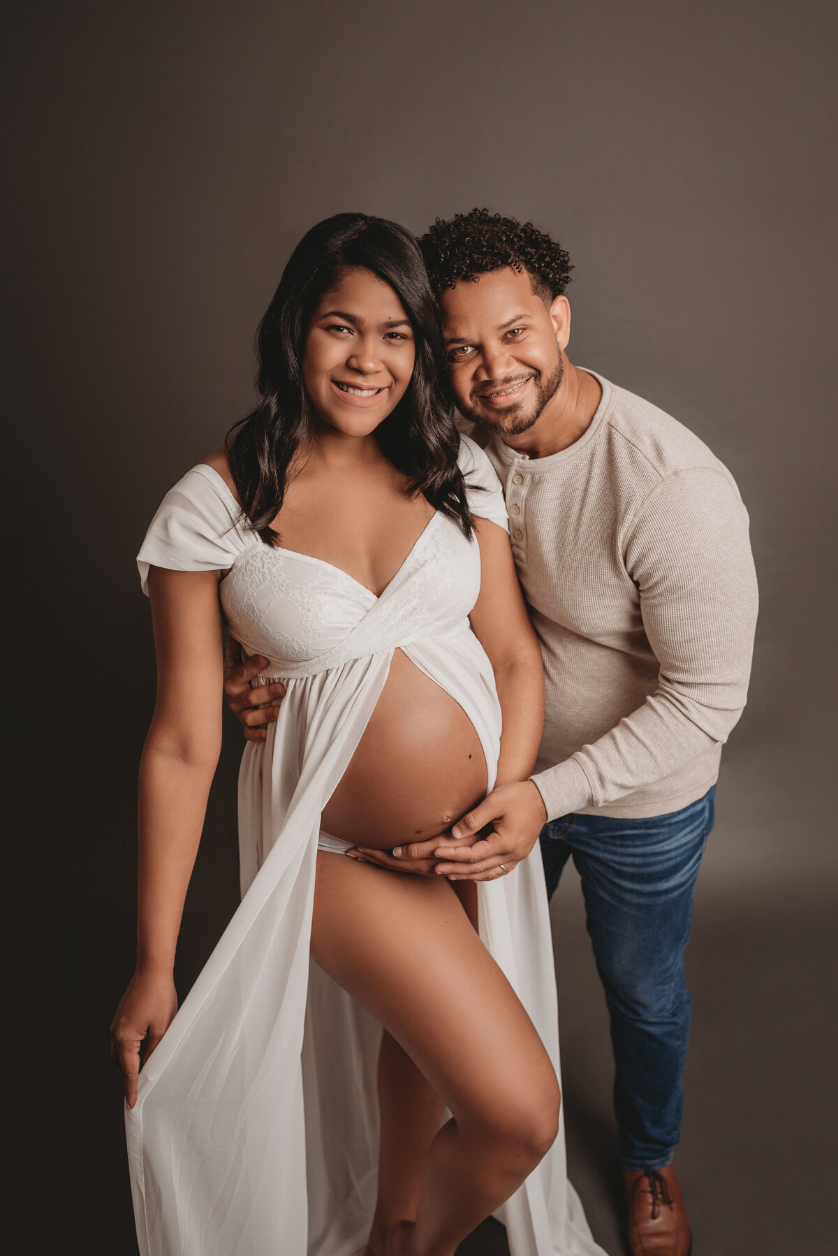 36 weeks pregnant couple woman showing bare belly and she and husband are holding her tummy. Woman is wearing white sheer maternity dress and man is wearing a cream long sleeve shirt with jeans and brown shoes.