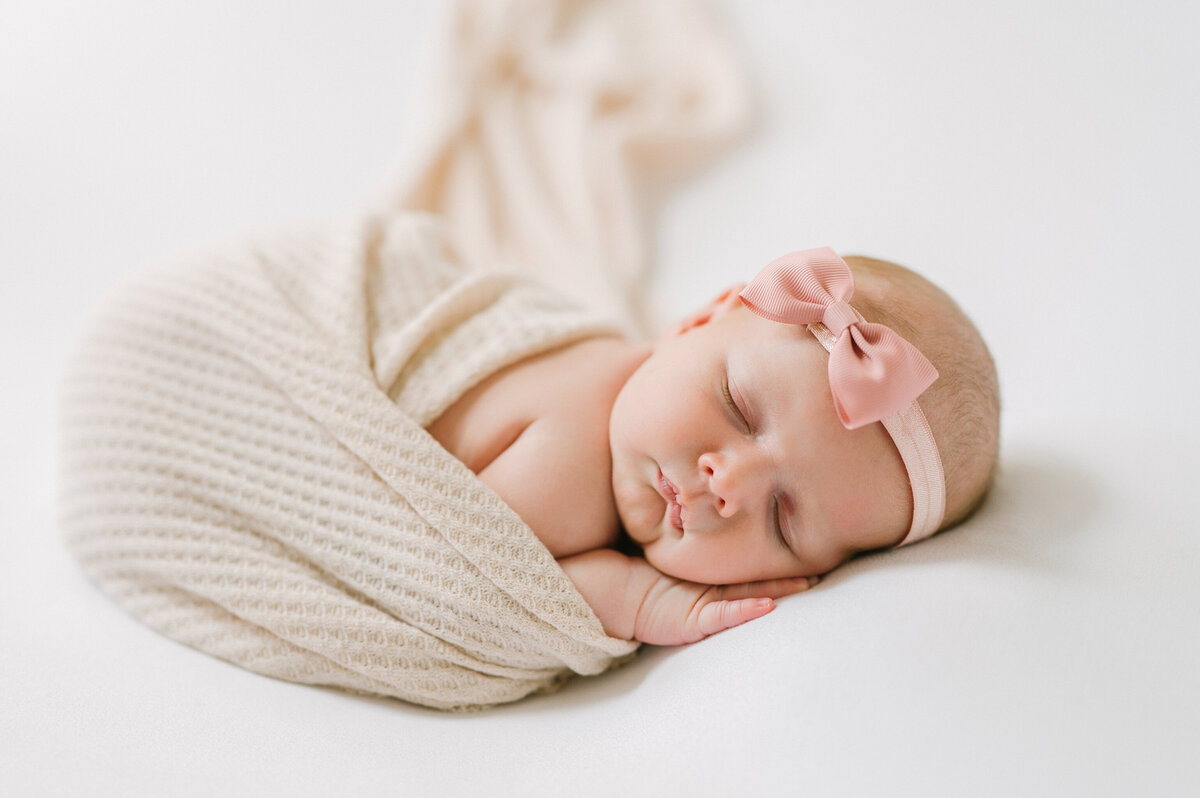 Newborn girl currled up on tummy wearing a pink bow and swaddle.