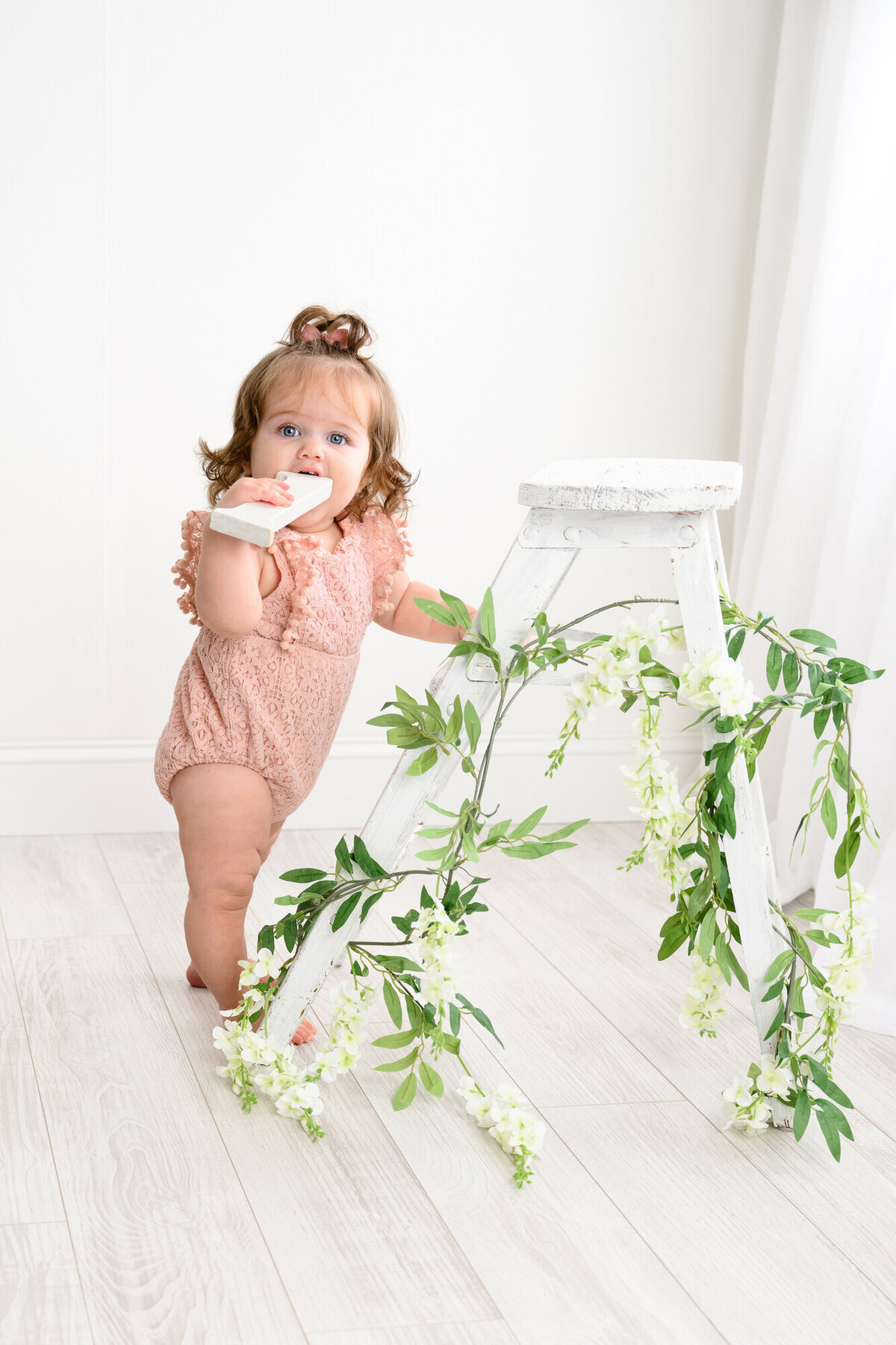 Charlotte Area 1-year-old stands next to a ladder covered in Ivy in a studio session wearing a pink romper and holding a number one prop