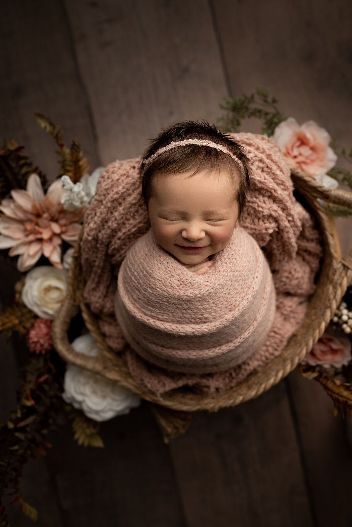 Fine art newborn photos captured by best Philadelphia newborn photographer Katie Marshall. Baby girl is swaddled in a pink light knit blanket and wearing a matching delicate headband. She is placed in a basket and there are coordinating flowers surrounding her.