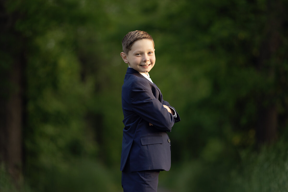 A young boy stands with arms crossed in a black suit smiling over his shoulder