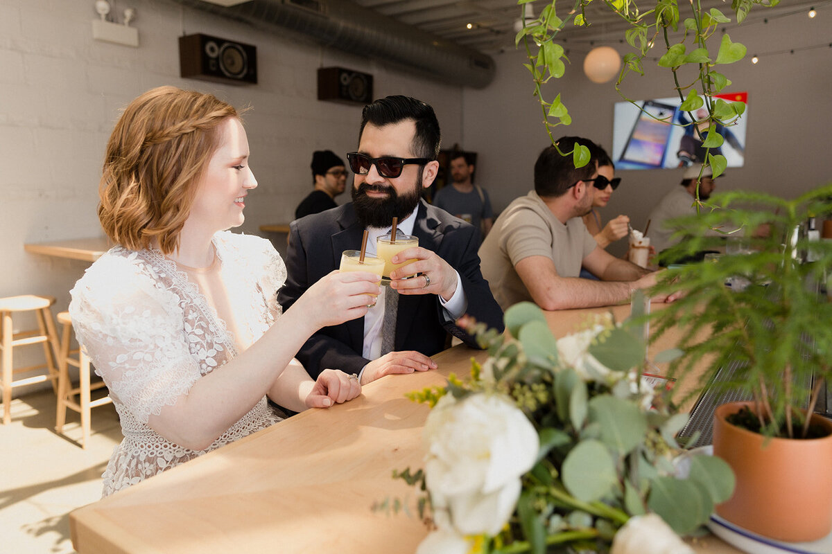 Wedding couple sitting at an outside bar on a sunny day. Groom wears sunglasses and they toast one another with drinks.