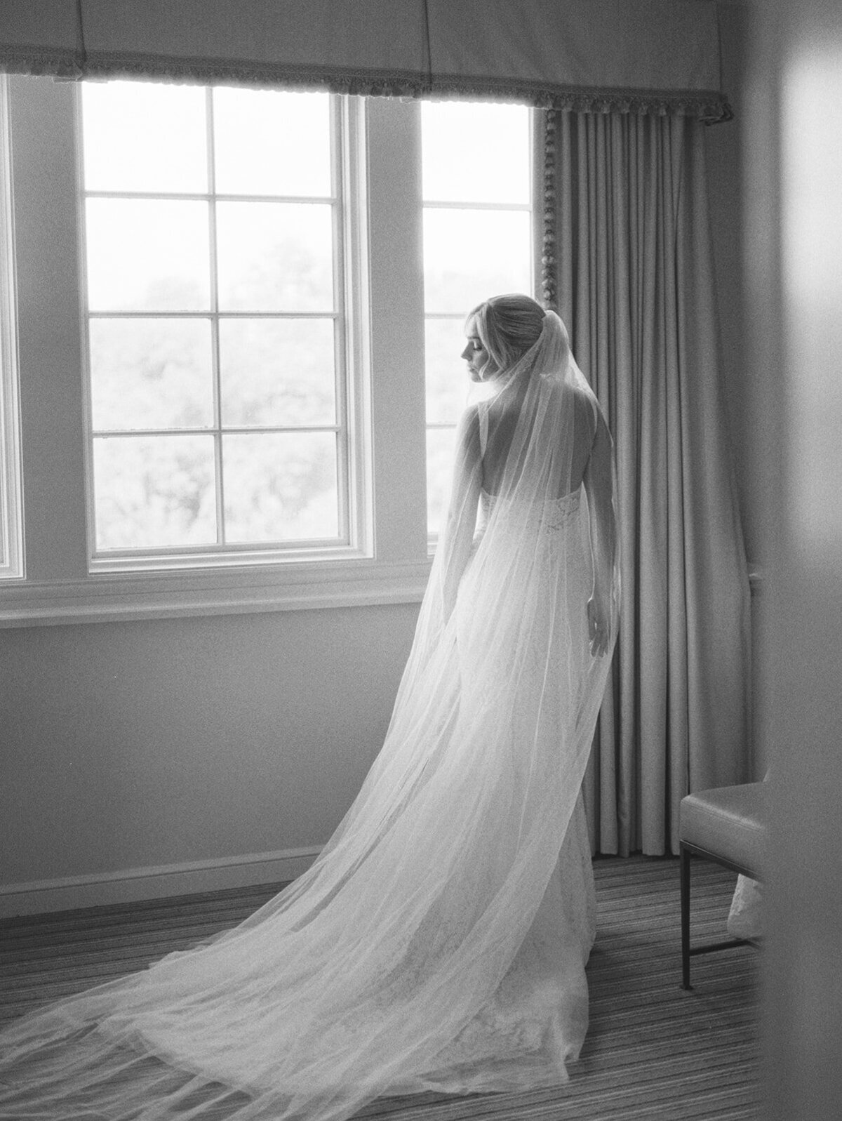 Bride looks over her shoulder as her veil is draped behind her  and the light illuminates her in this black and white film image