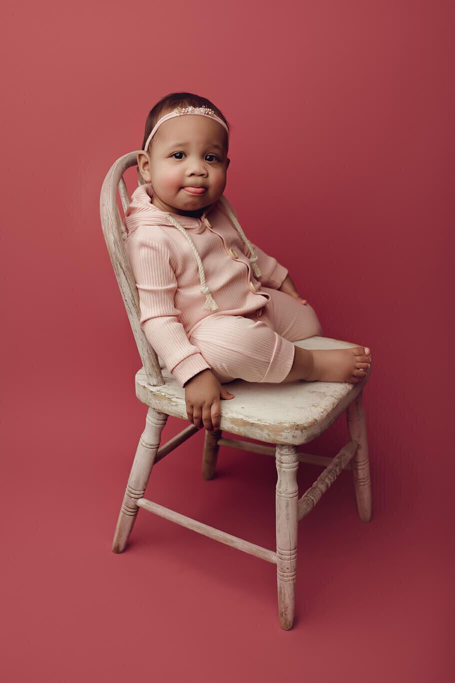 a one year old girl sitting on a white chair on a pink backdrop pouting her bottom lip