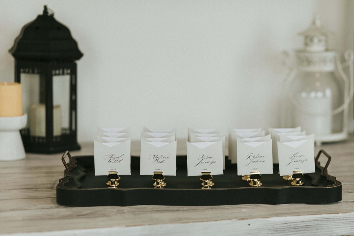 White wedding place cards with black cursive font on gold place card holders atop a tray with lanterns in the background.