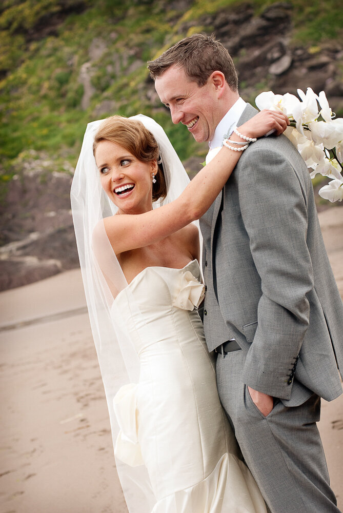 brunette bride wearing a mermaid style wedding dress, holding a white flower bouquet hugging her husband who is wearing a light grey suit with waistcoat, standing on the beach in Kerry