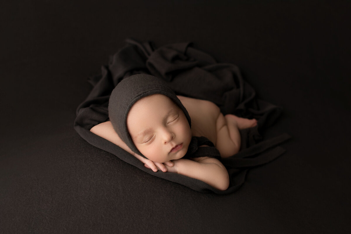 Fine art newborn photos captured by best Main Line newborn photographer Katie Marshall. Baby boy is sleeping on his belly with his fands folded under his chin. his head is resting on his forearm. Baby is wearing a brown bonnet and laying atop of brown stretch fabric.