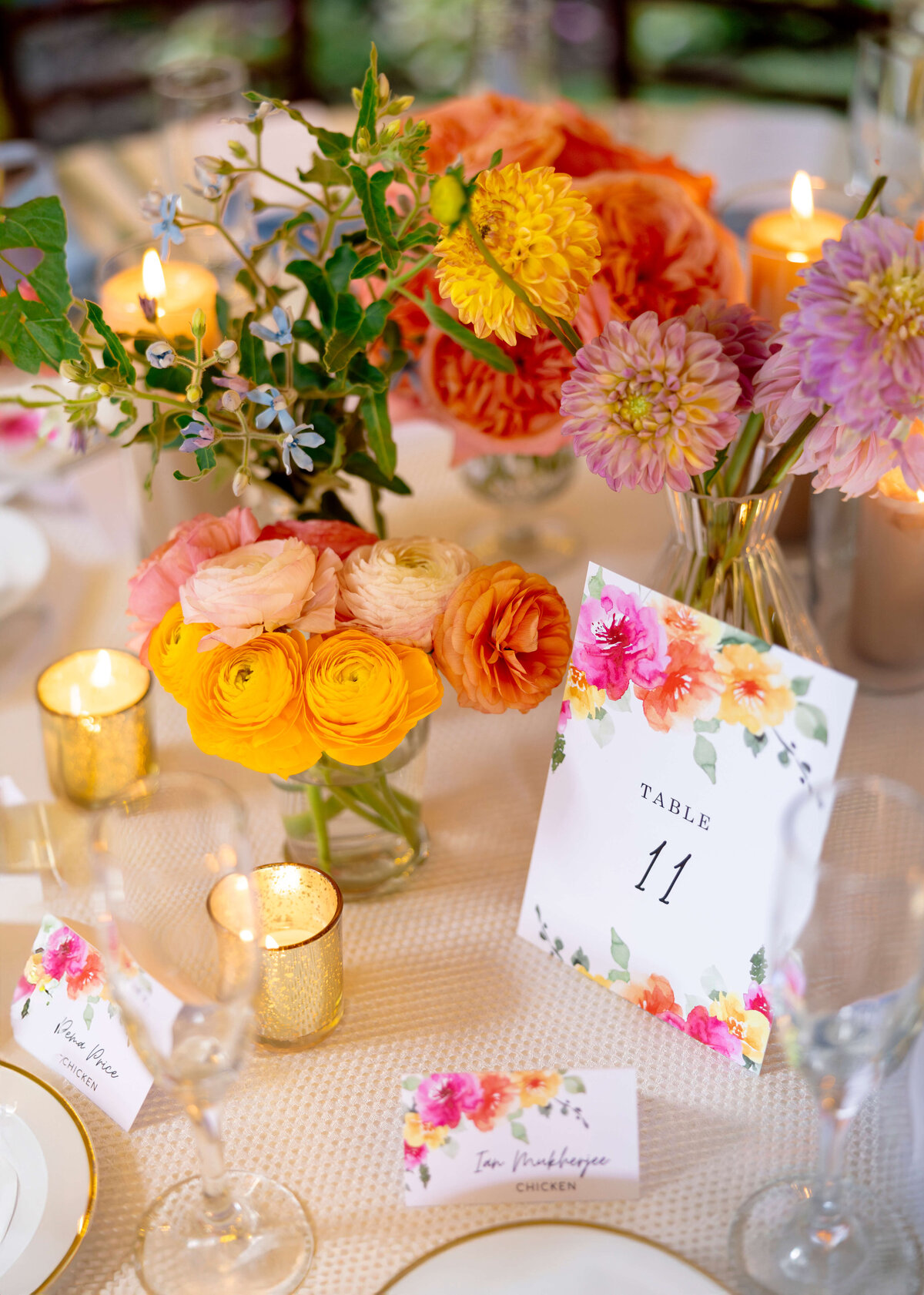 A beautifully arranged table features vibrant floral centerpieces with orange, pink, and yellow blooms. Lit candles in gold holders add warmth. A card displaying Table 11 and menus with floral designs complement the elegant setting.