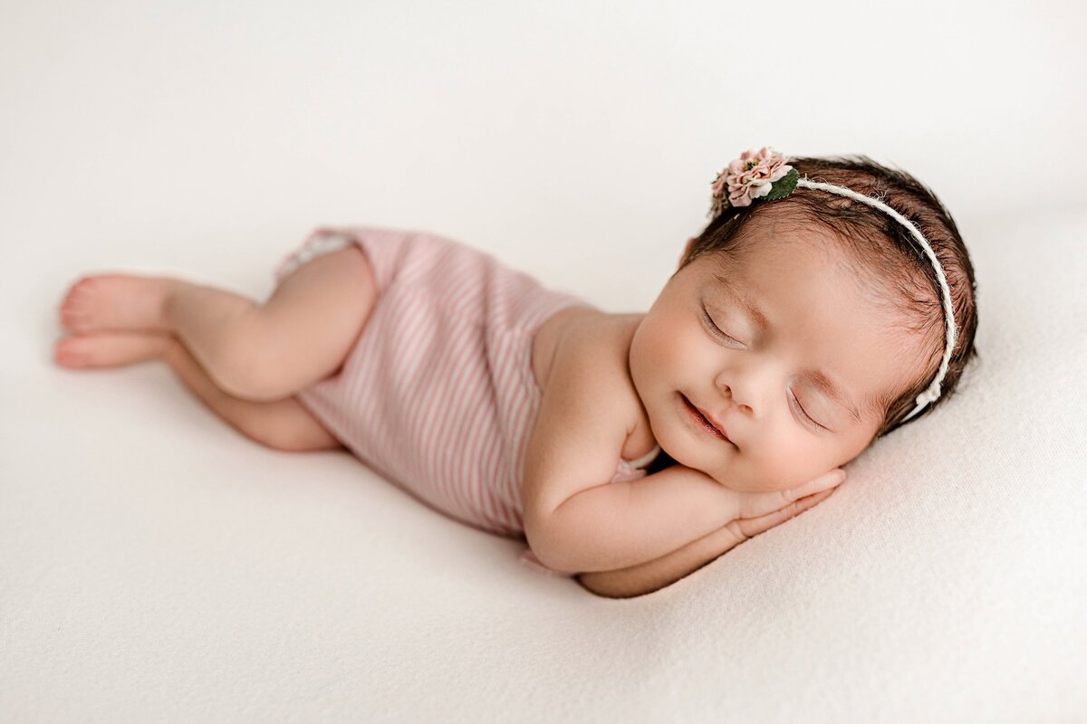  Baby girl dressed in a pink striped outfit lays on her side with her hands underneath her cheek wearing a pink flower headband during her Studio newborn session